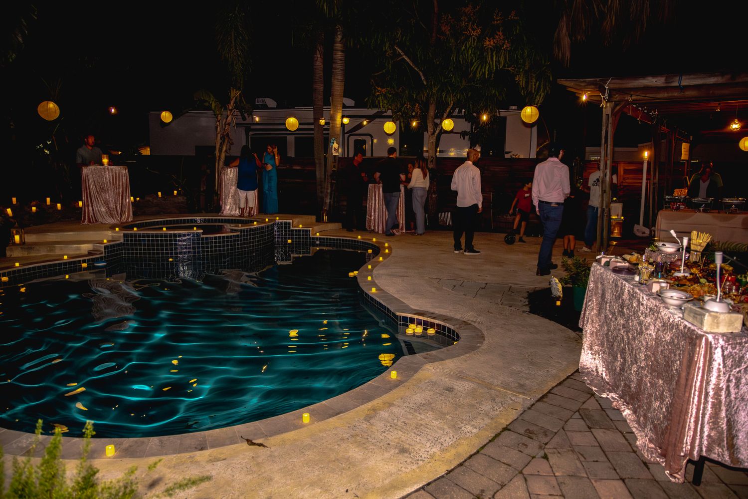 Illuminated swimming pool at night with blue lights reflecting on water and guests gathered around a decorated buffet table.