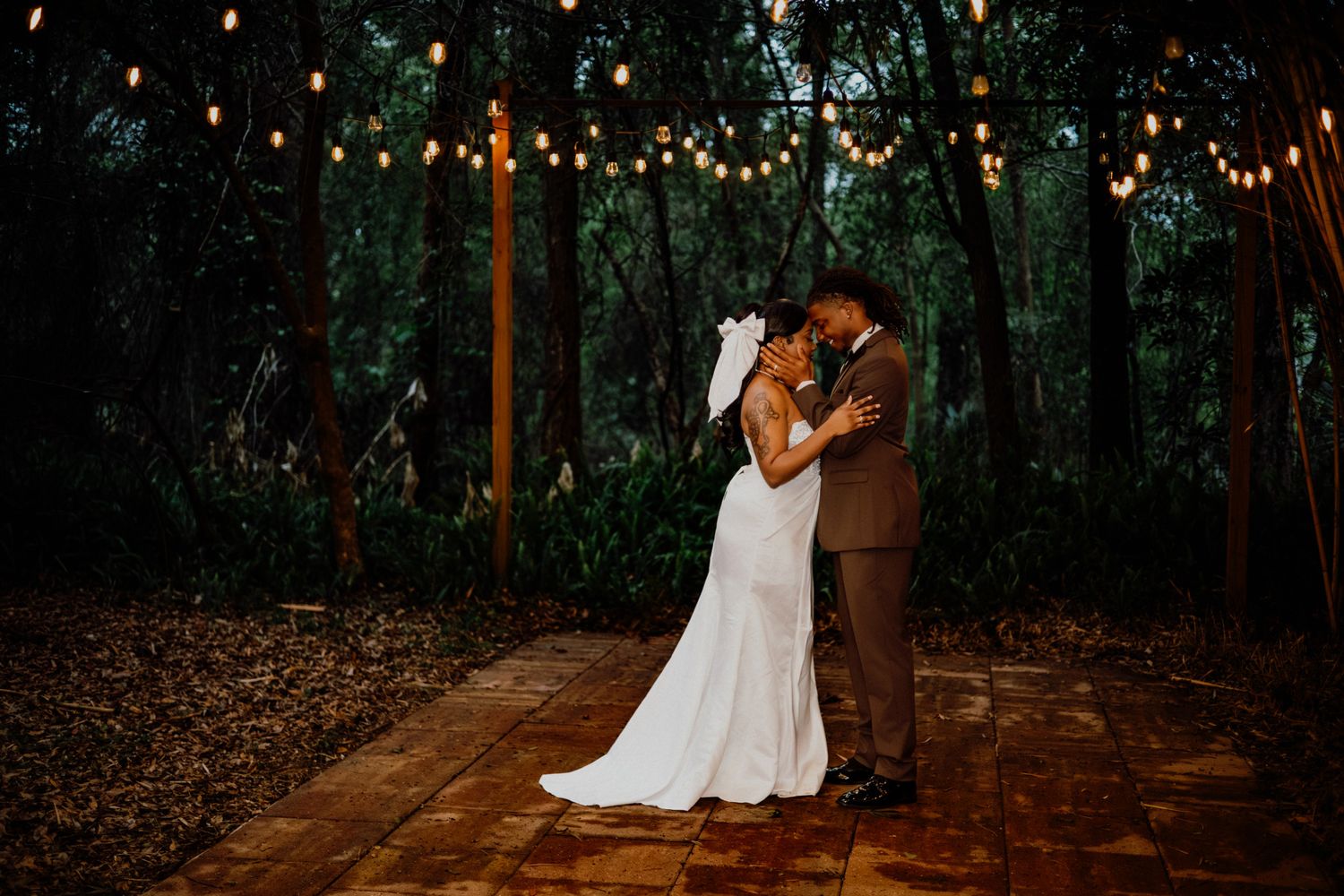 A romantic outdoor wedding ceremony takes place on a wooden deck illuminated by warm string lights at dusk.