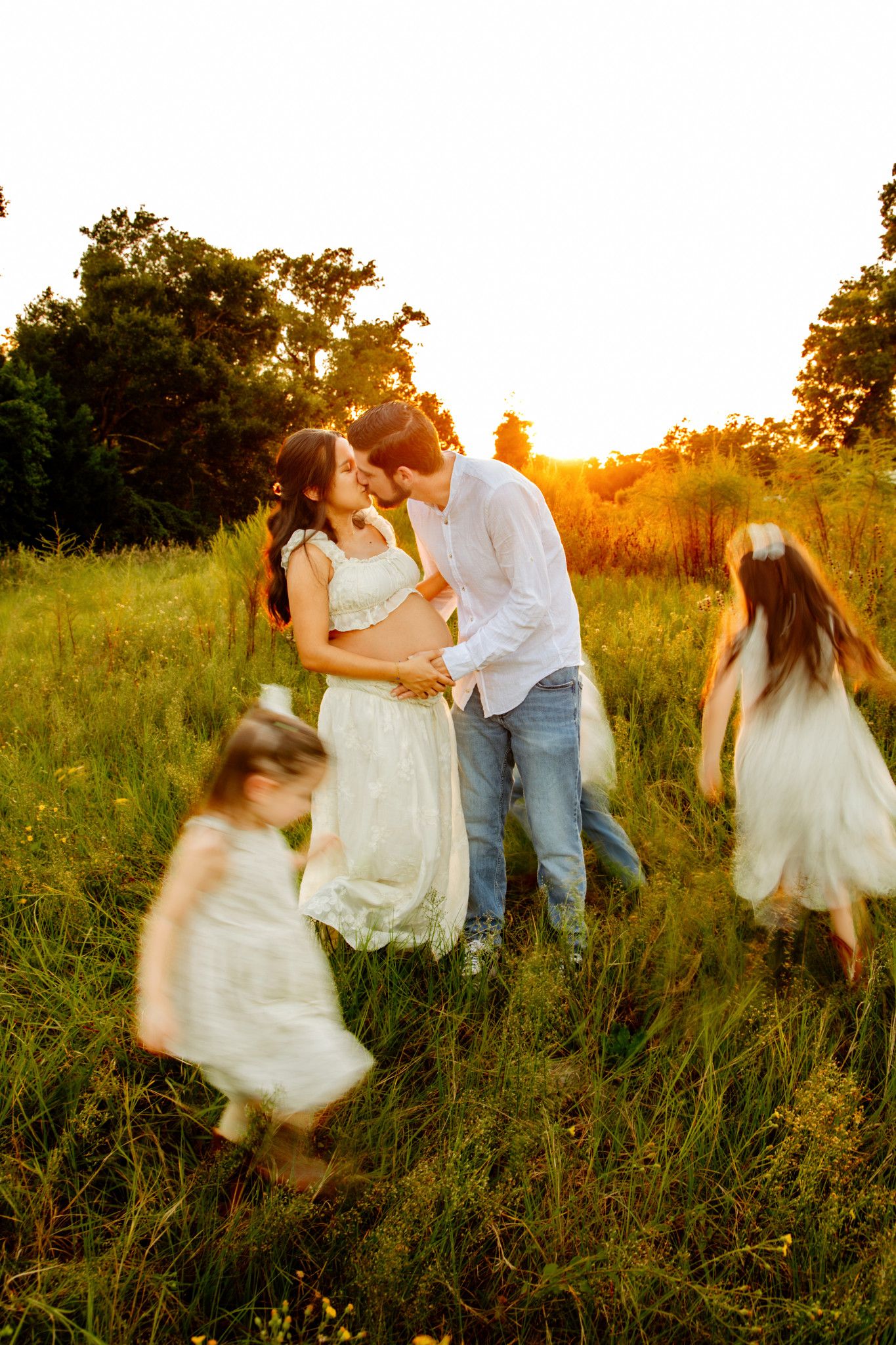 Couple kissing at golden hour in field while two girls in white dresses twirl around them.