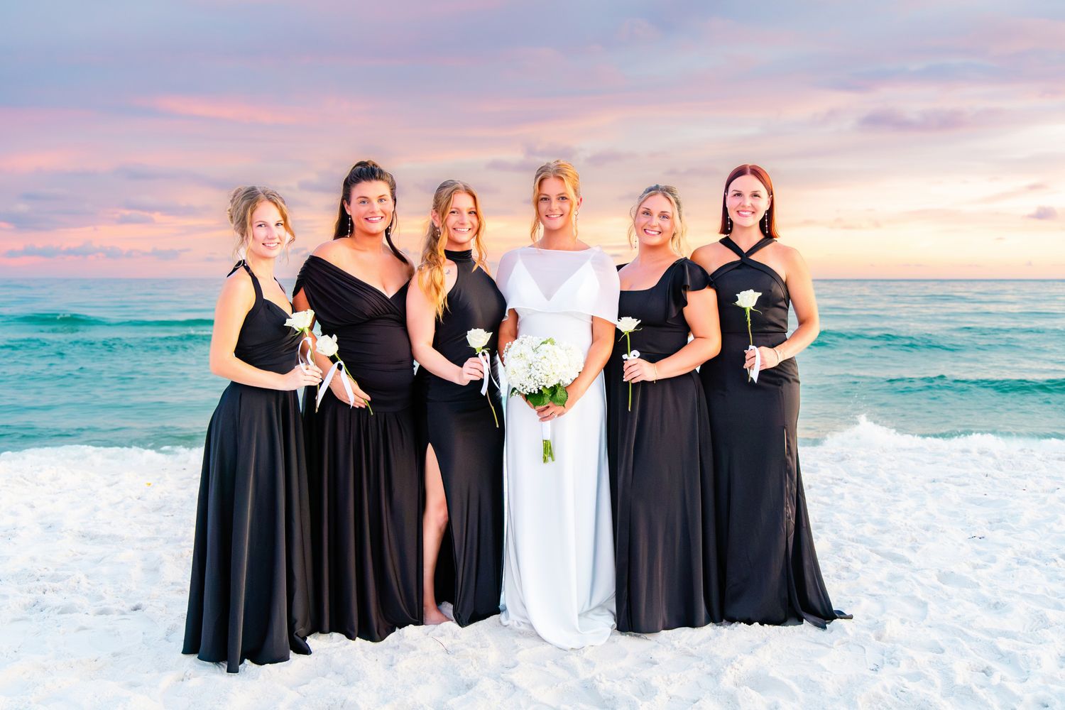 Bride in white gown with bridesmaids in black dresses on a sandy beach at sunset.