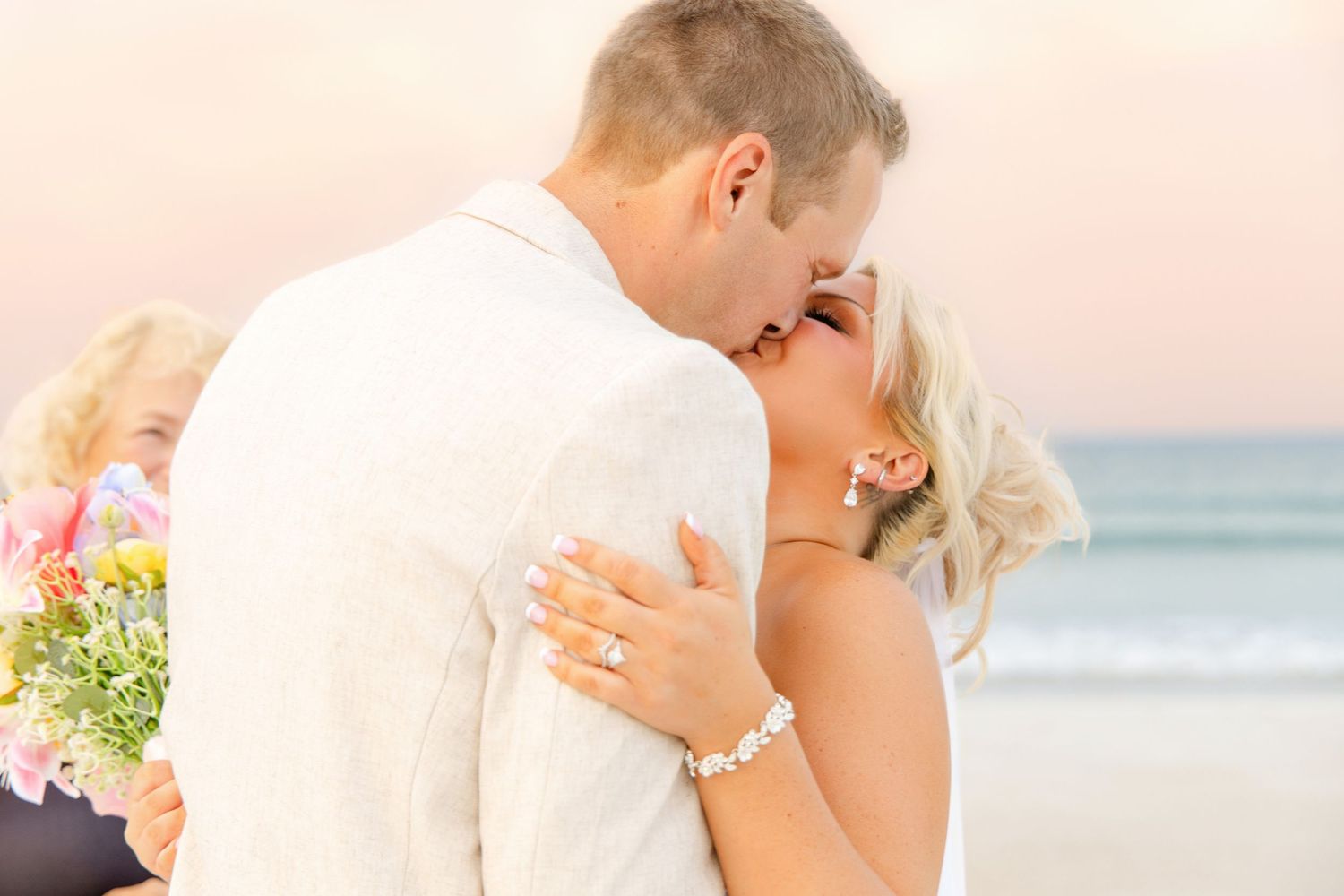 Couple sharing a romantic kiss on the beach during their wedding ceremony, bride wearing a white dress and pearl bracelet.