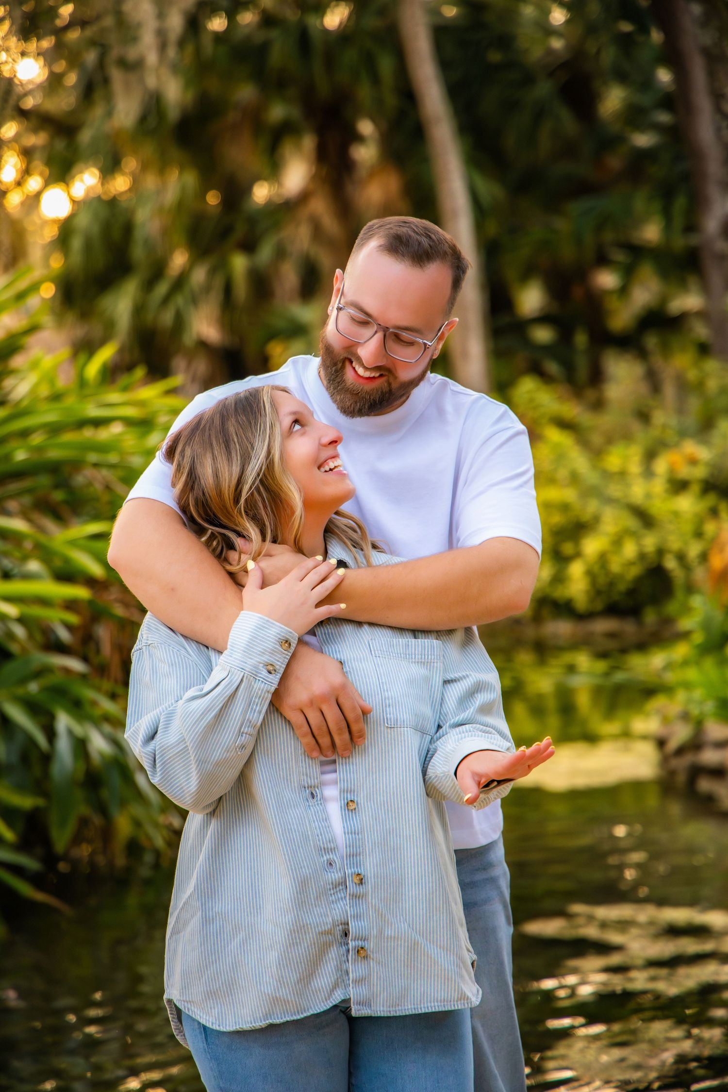 Couple in casual attire embraces lovingly in a lush garden setting with golden sunlight filtering through trees.