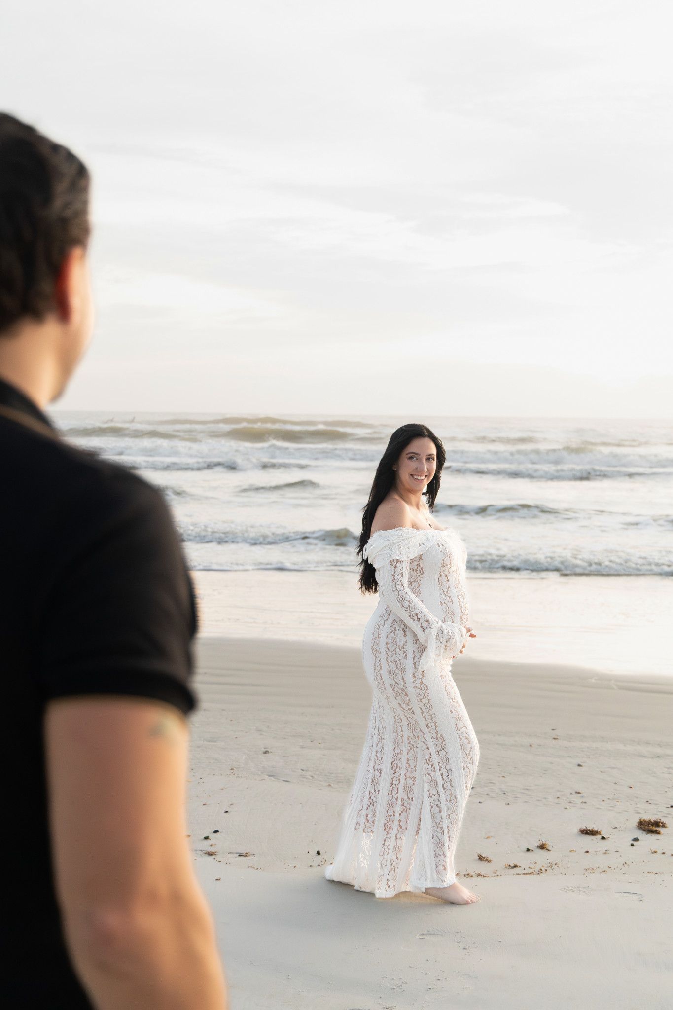 Pregnant woman in white lace maternity gown smiles at man on sandy beach with ocean waves behind her.
