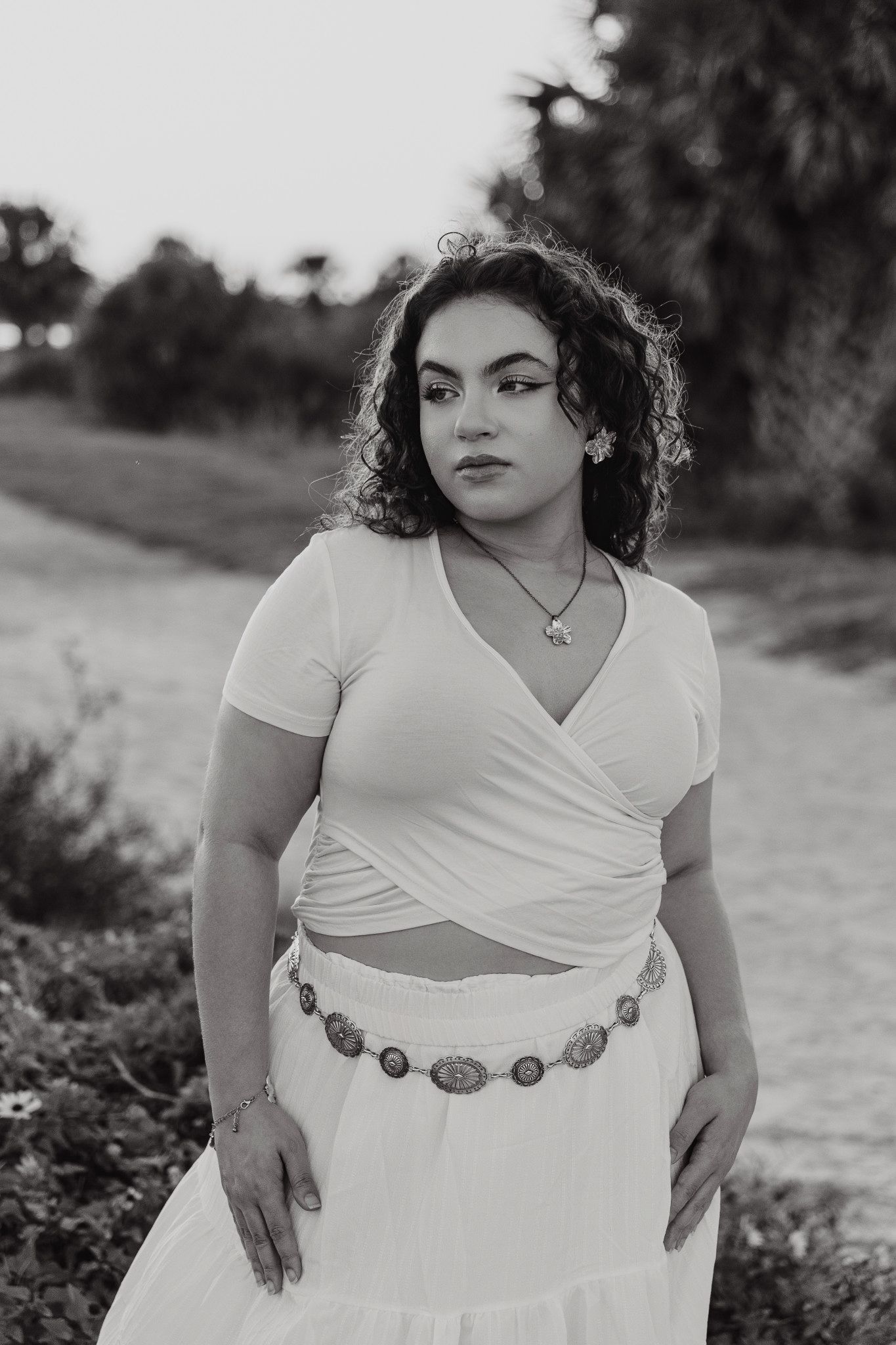 Black and white portrait of a woman with curly hair in a white wrap top and skirt with a concho belt standing outdoors by a coastal path.