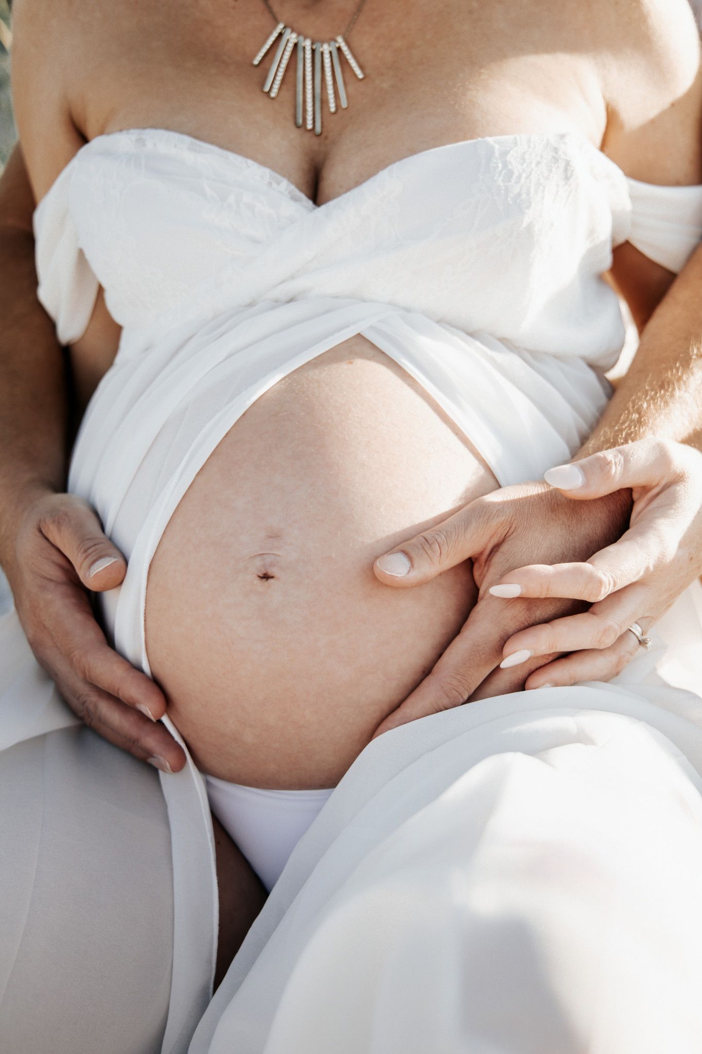 Pregnant woman in white dress with partner's hands cradling her bare baby bump, wearing a silver necklace.