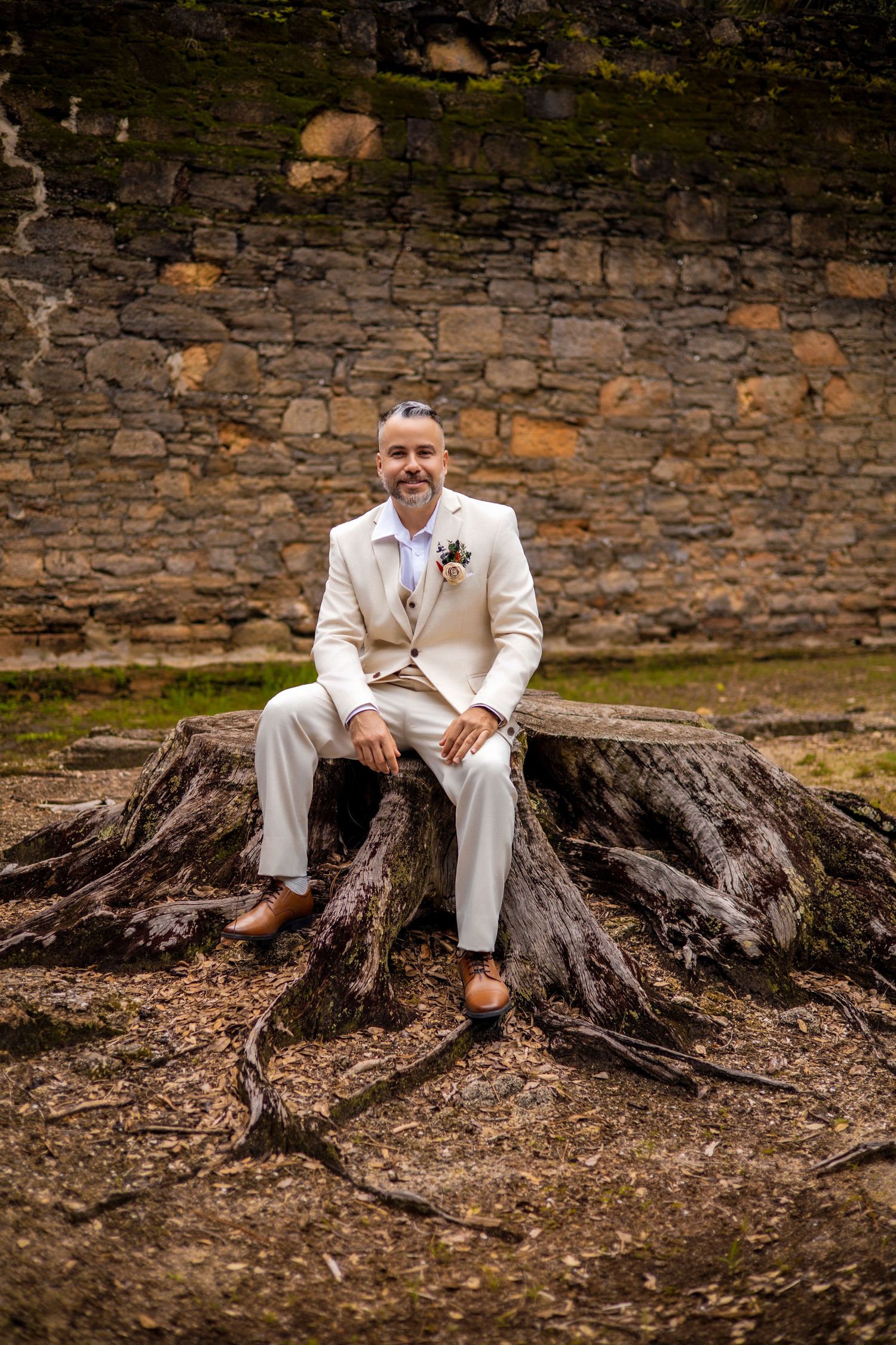 A person in a cream suit sits casually on large tree roots against a stone wall backdrop.