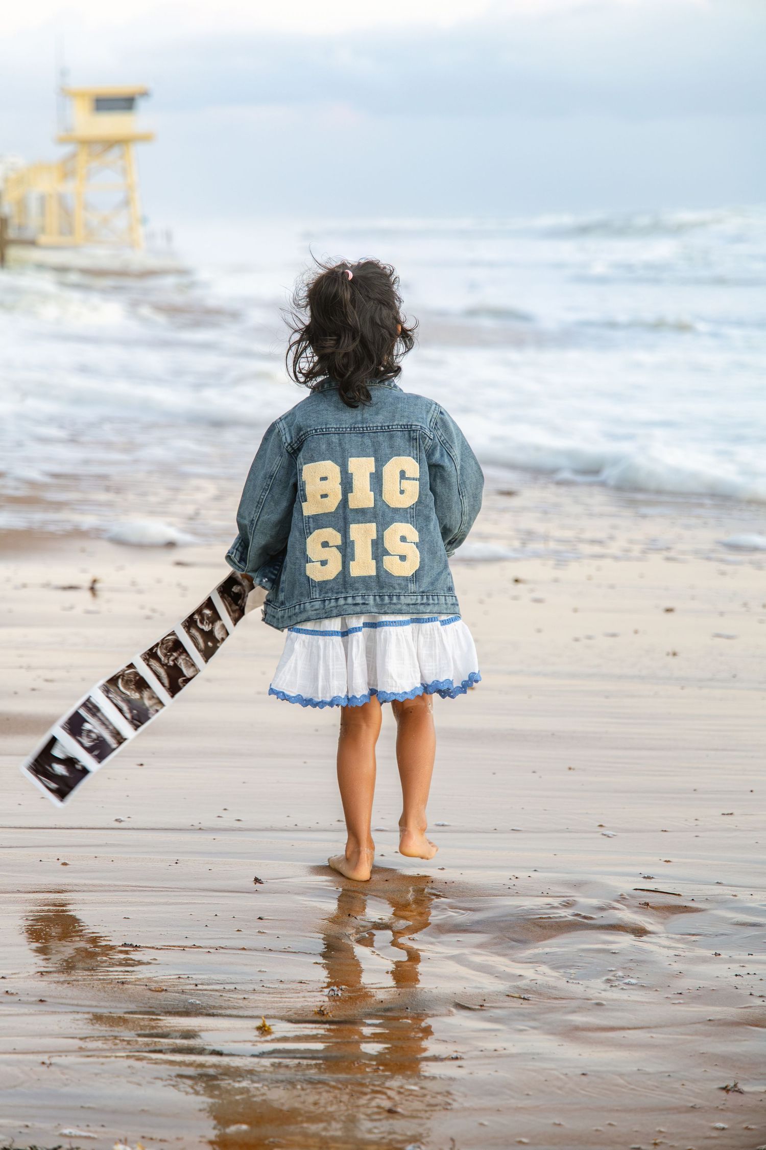 Young girl in a denim jacket reading BIG SIS walks barefoot on the beach holding an ultrasound photo strip near the ocean waves.