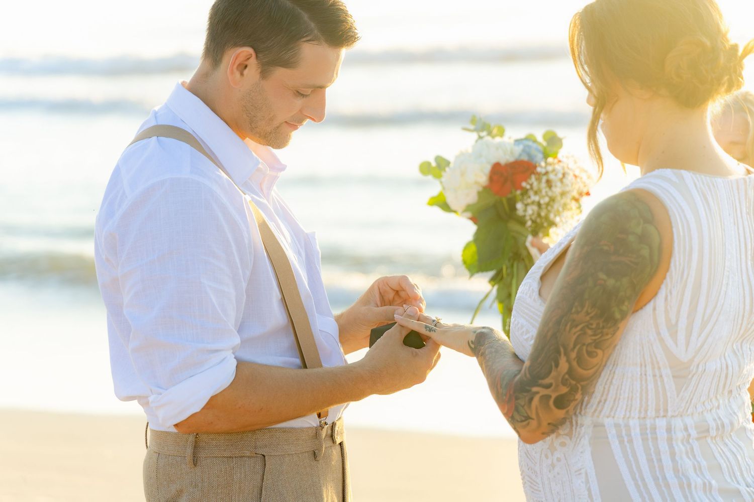 Groom placing wedding ring on bride's finger during beach ceremony at golden sunset.