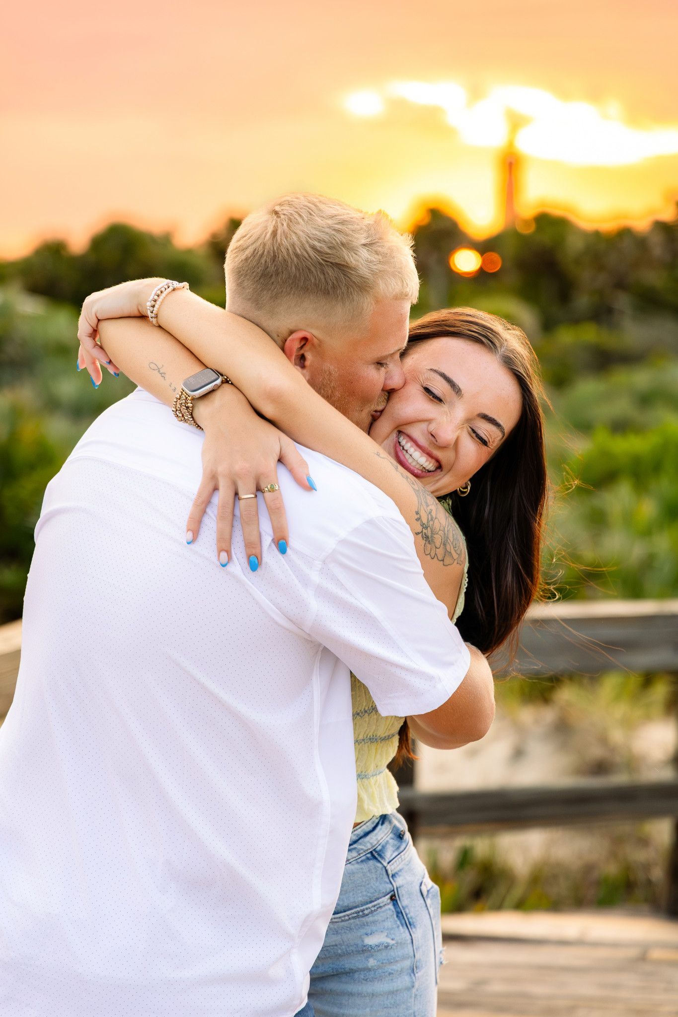 Couple embracing at sunset on a boardwalk, woman smiling as man kisses her cheek with golden sky behind.