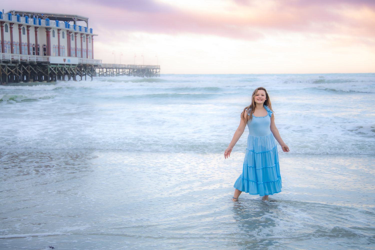 Young woman in blue dress wading in ocean waves at sunset near a pier.