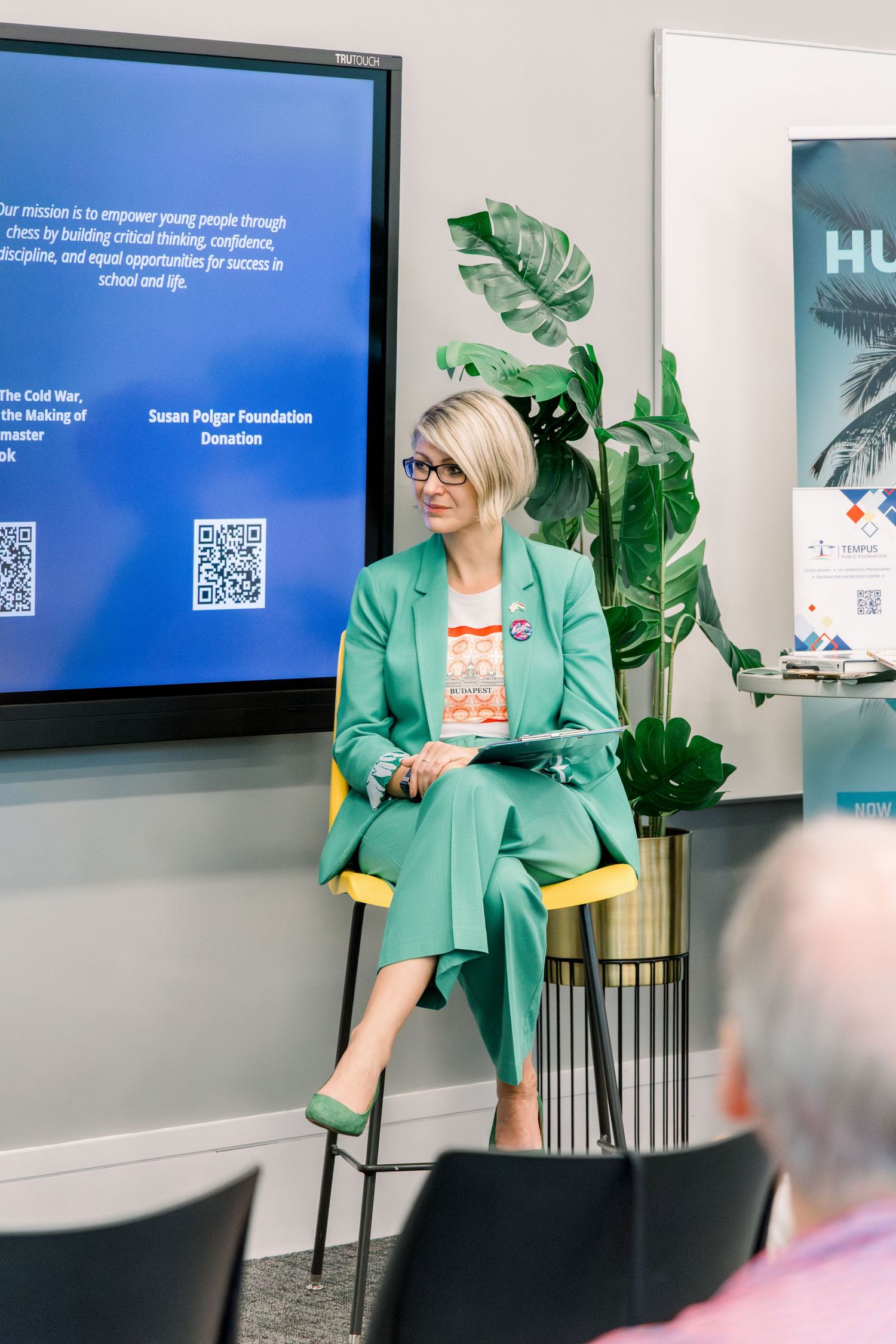 Woman in green suit sits on chair presenting at a professional event with a blue screen displaying a QR code.