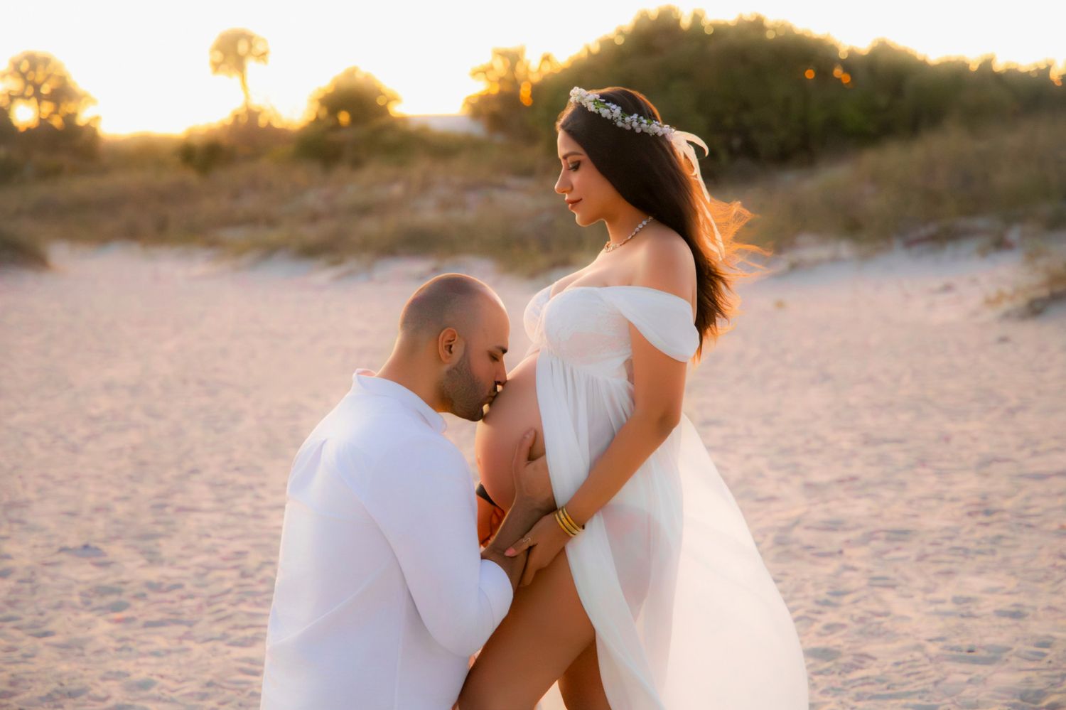 Couple in white outfits sharing a tender maternity moment on a sandy beach at golden sunset.