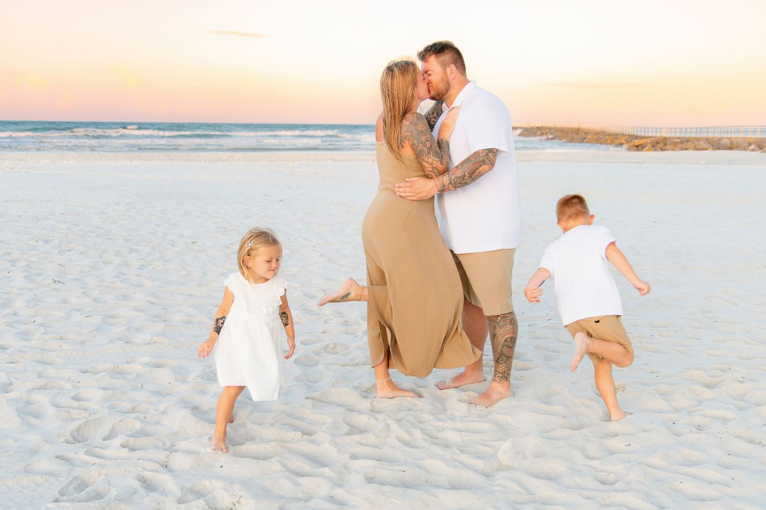 Family beach photo at sunset with couple kissing and two young children playing on white sand.