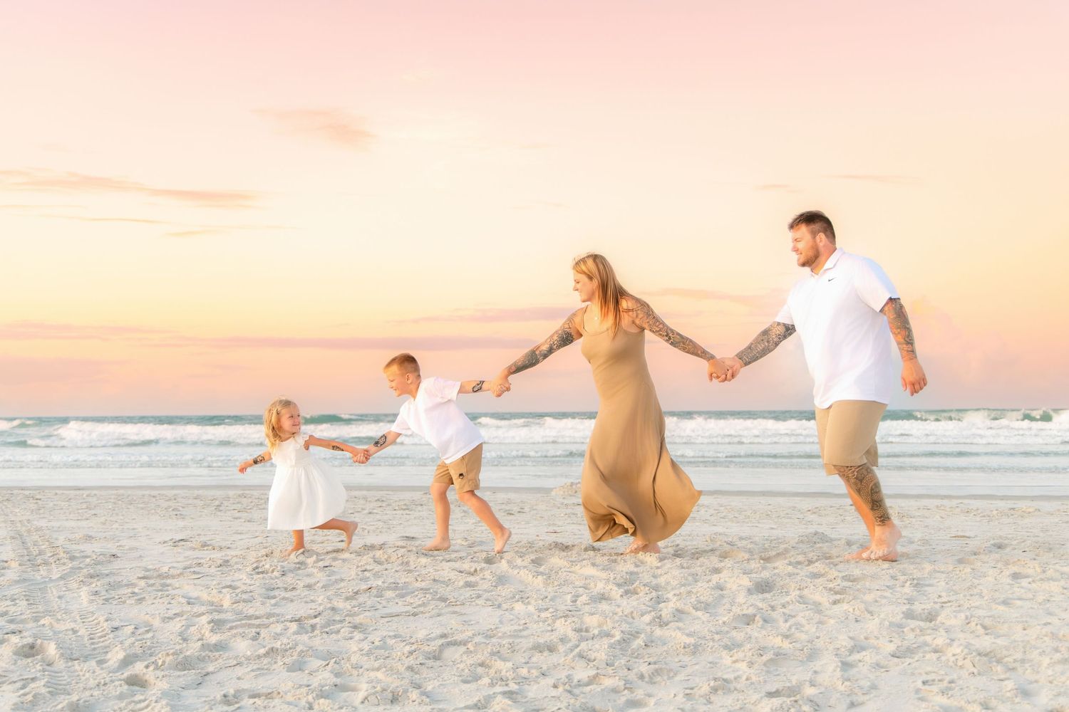Family of four holding hands and playing on a sandy beach at sunset with pastel pink and orange skies.