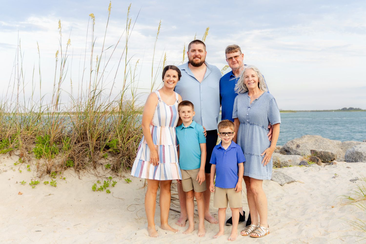 Family of six posing together on a sandy beach near rocks and sea grass with calm ocean water in the background.