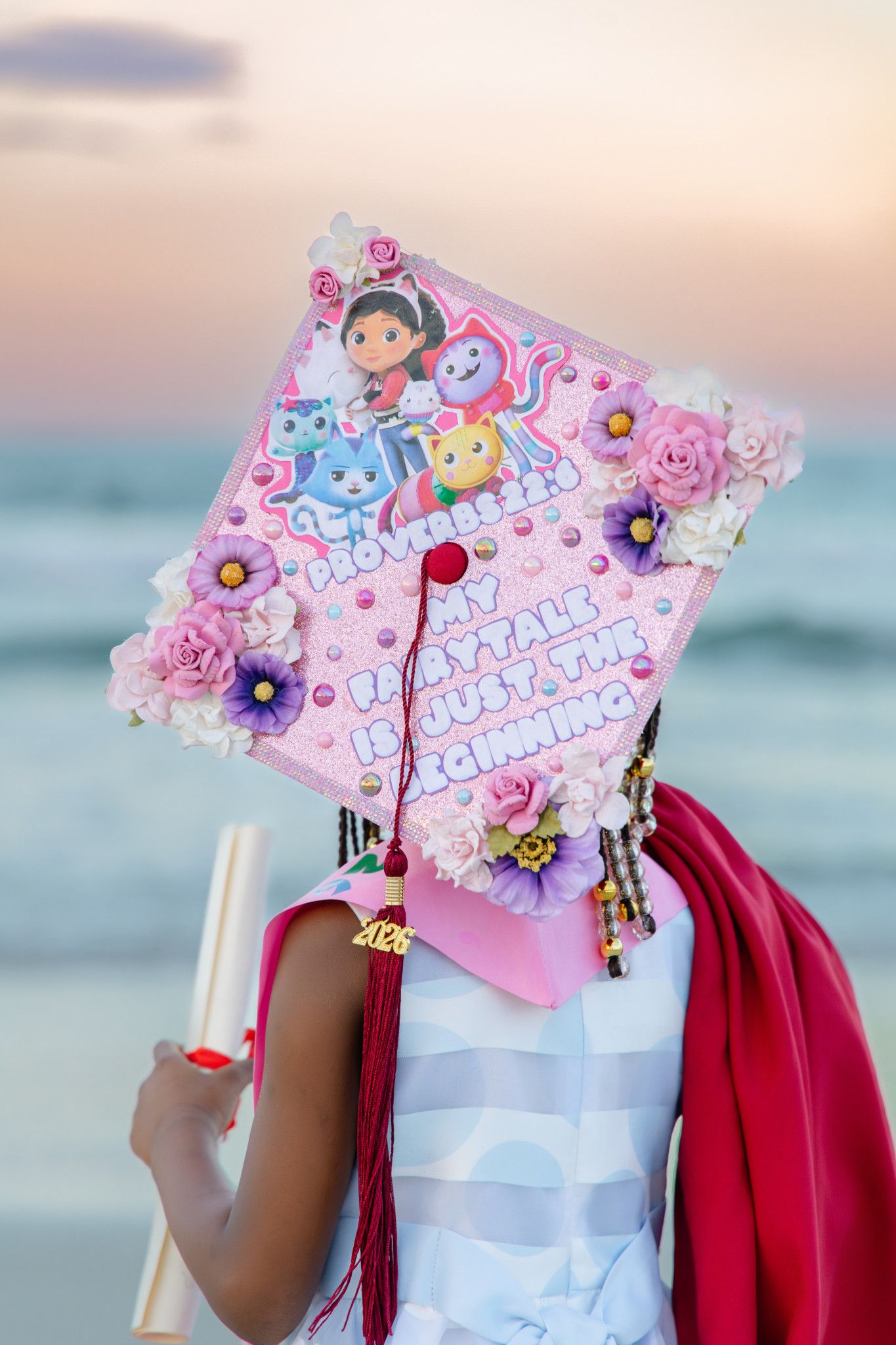 Graduate holds diploma at beach sunset, wearing decorated pink Gabby's Dollhouse graduation cap with flowers and pearls.