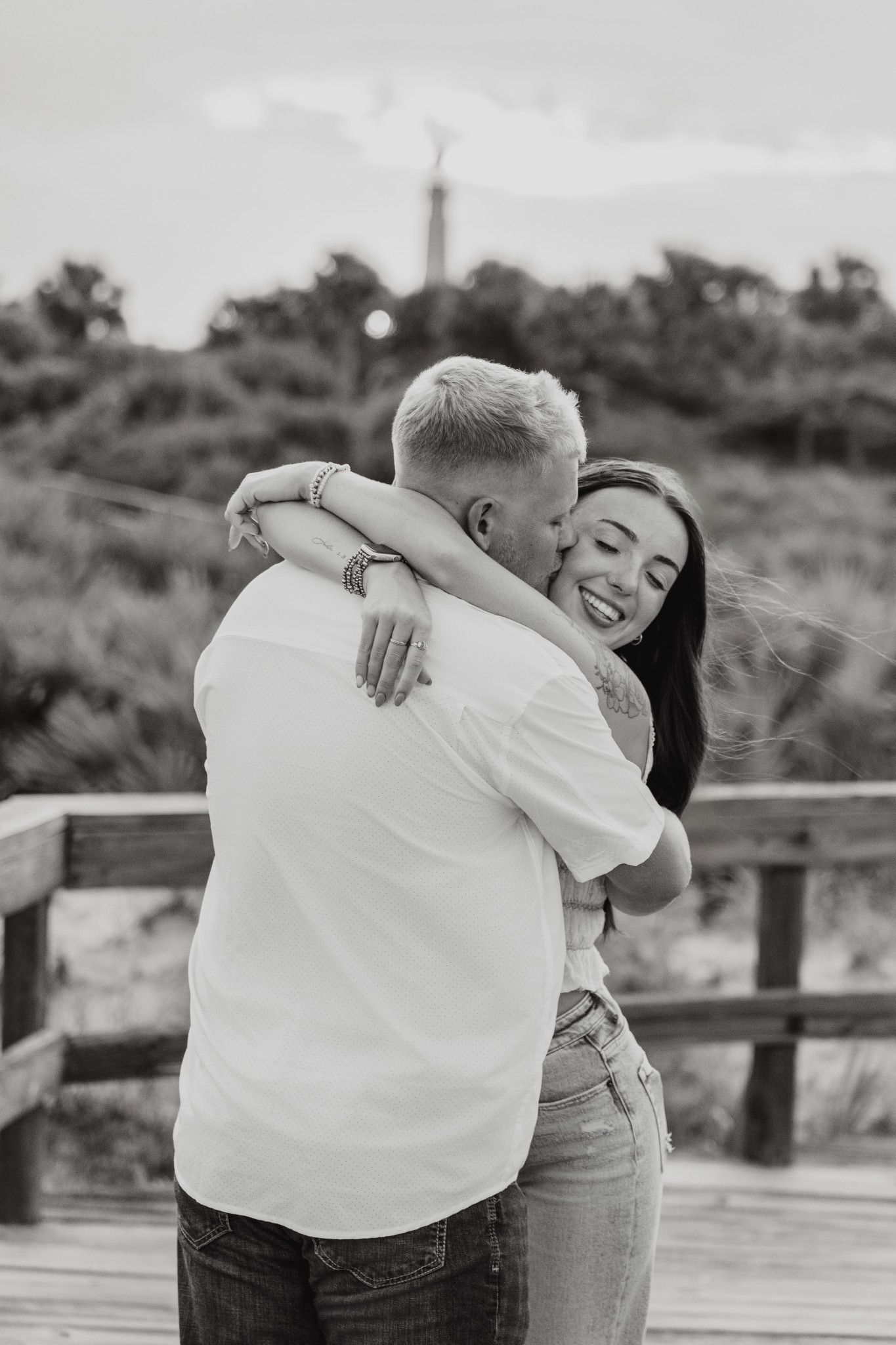 Black and white photo of a smiling couple embracing on a wooden boardwalk with lush greenery in the background.