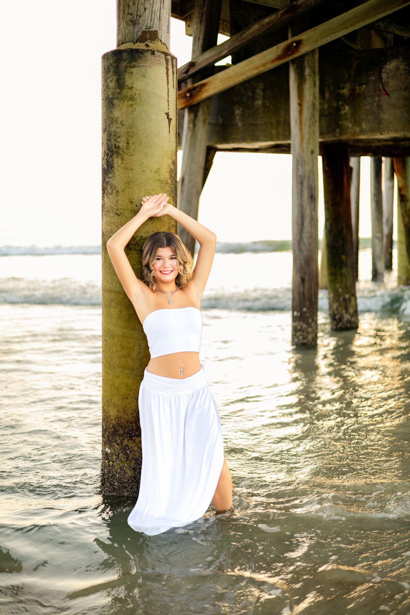 Young woman in white crop top and skirt posing under a pier in shallow ocean water with hands raised.