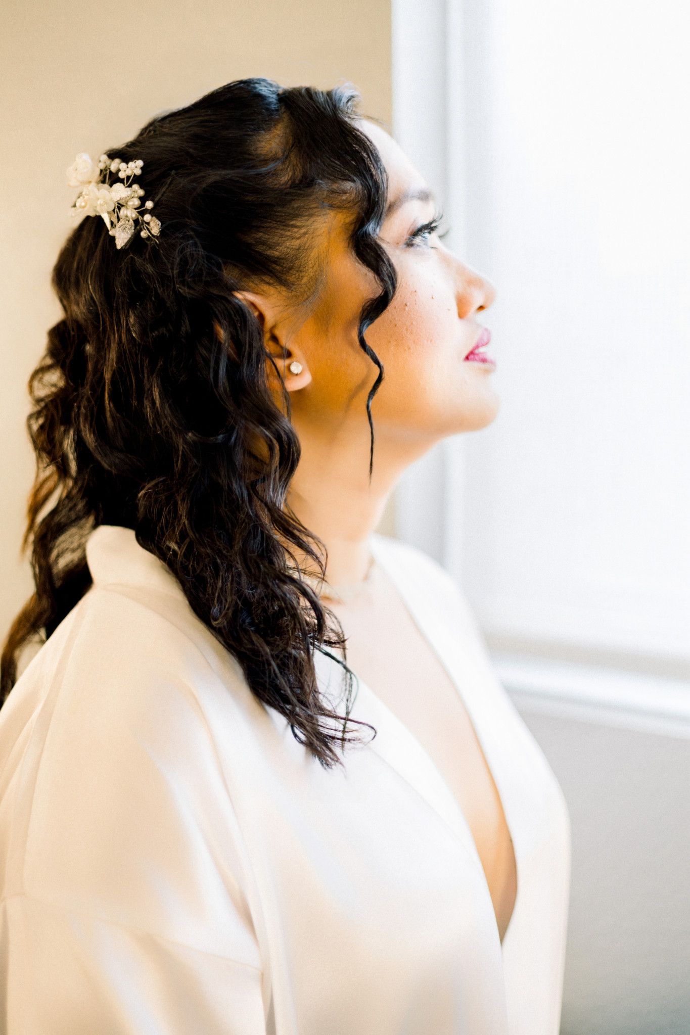 Bride in white robe with floral hair accessories gazing upward near a bright window on her wedding day.
