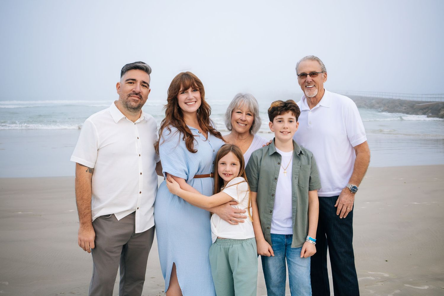 A happy family poses together on a sandy beach wearing coordinated blue and white outfits during a cloudy day.