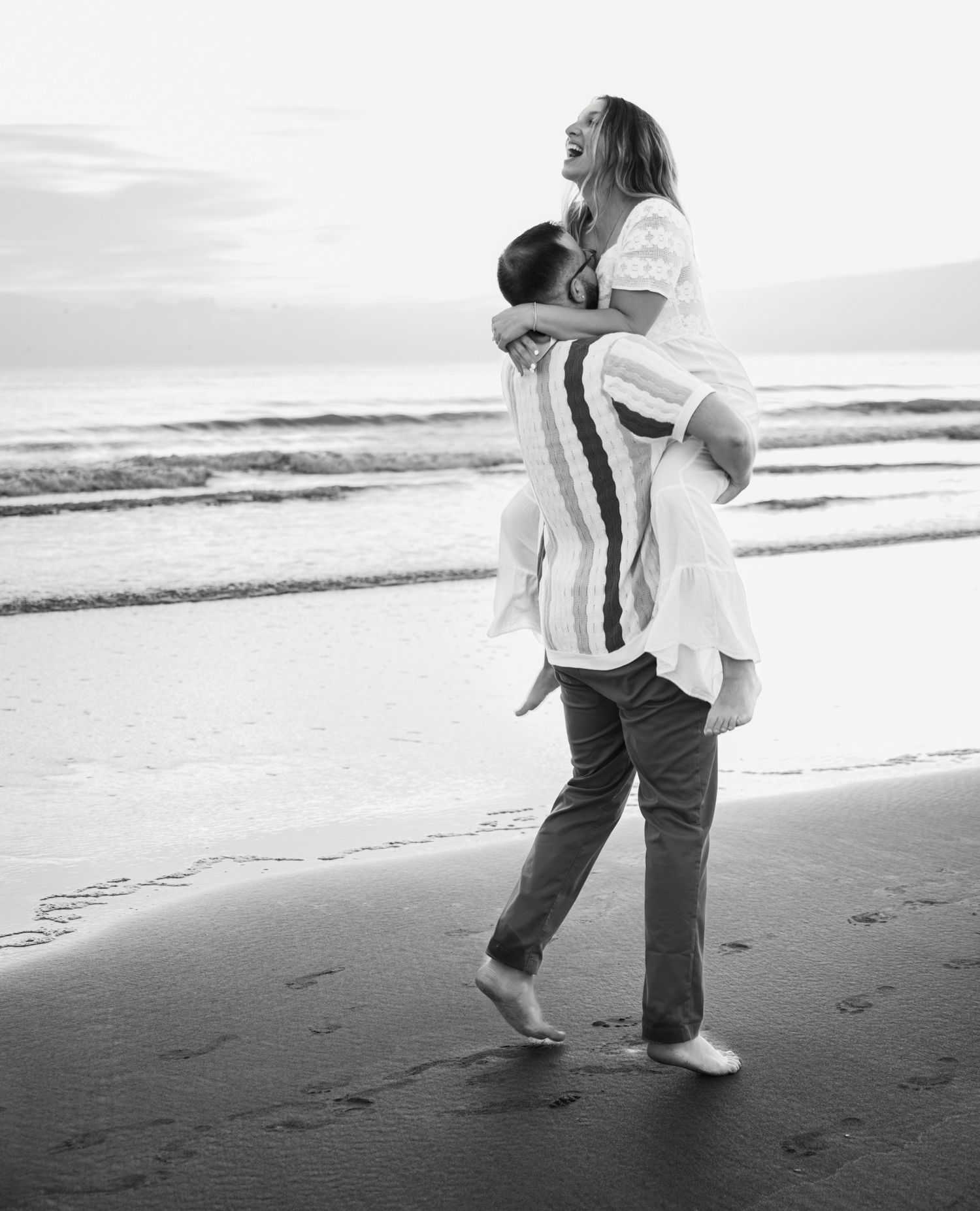 A couple in casual attire dances together on a serene beach at sunset, creating a romantic silhouette against the ocean.