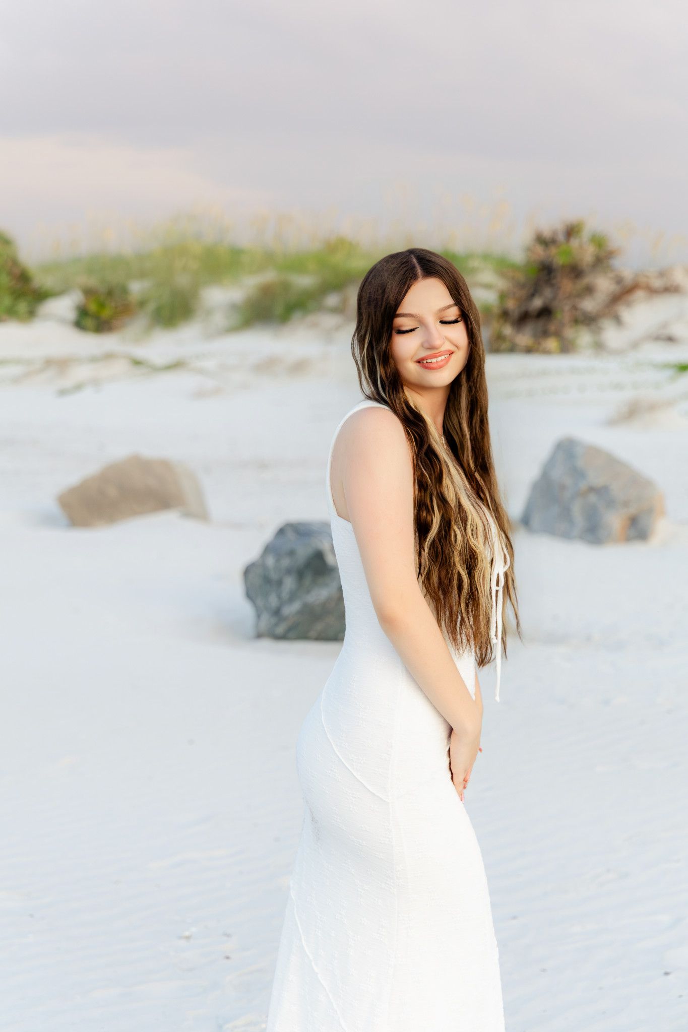 Young woman in white dress smiling on sandy beach with rocks and dunes at sunset.