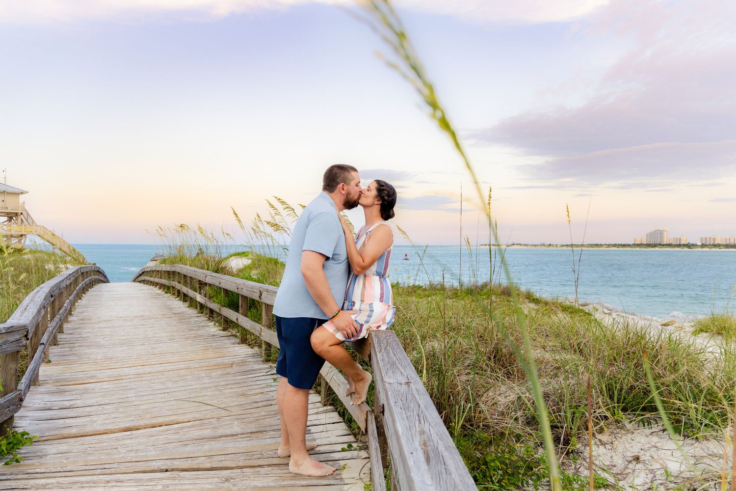 Couple kissing on a wooden boardwalk at sunset near a sandy beach with sea oats and turquoise water.