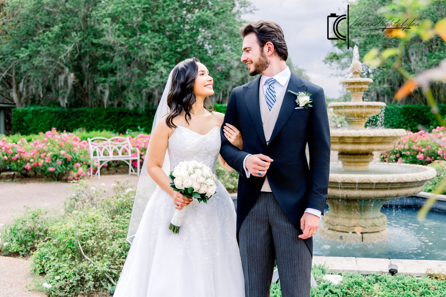 Bride and groom pose by a stone fountain in a garden, she holds white flowers, he wears a navy pinstripe suit.