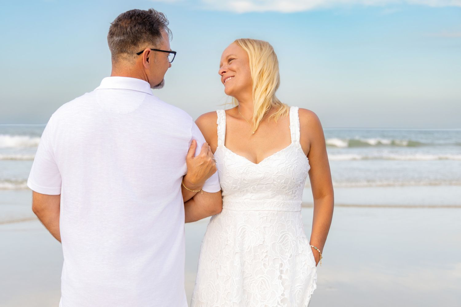 Couple in white outfits holding hands on a sunny beach, woman in lace dress smiling at man.