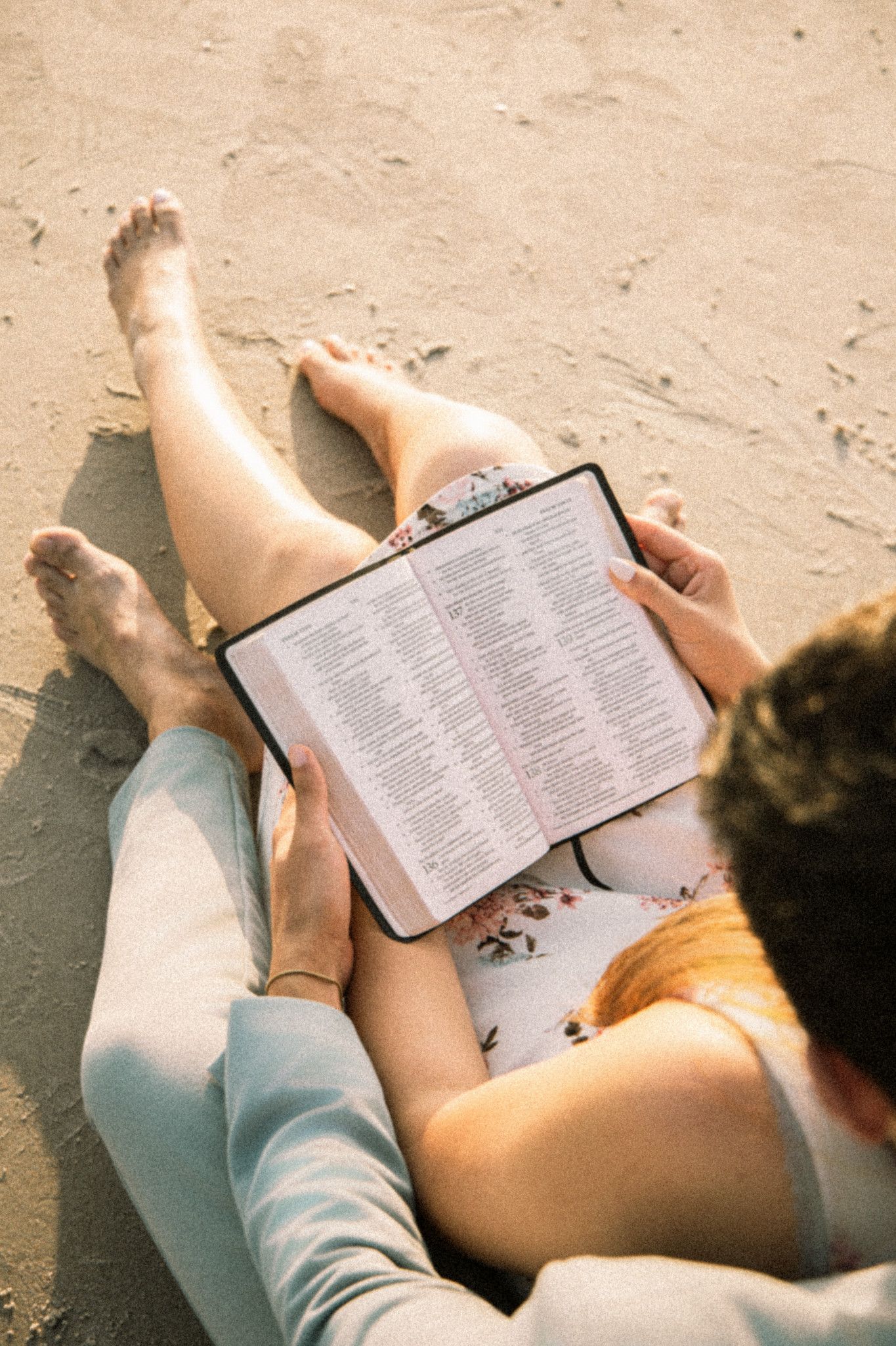 Couple sitting on sandy beach reading Bible together, barefoot with legs intertwined, sharing a peaceful spiritual moment outdoors.