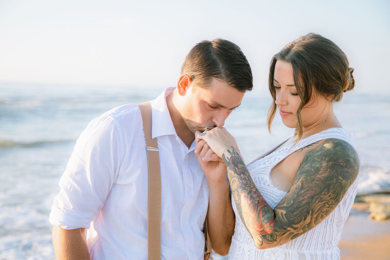 Groom kisses tattooed bride's hand tenderly on a sunny beach during wedding ceremony.