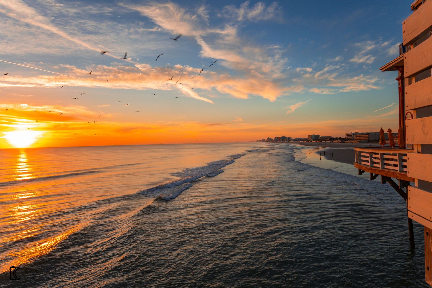 Panoramic view of a vibrant sunset over the ocean with golden clouds reflecting on calm waters along a coastal pier.