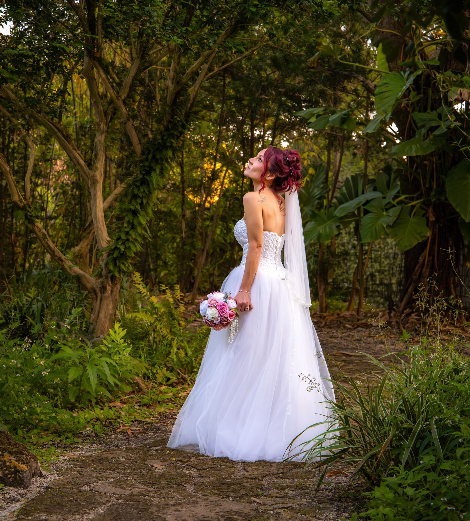 A bride in a white wedding dress walks along a wooded path holding a pink and white bouquet.