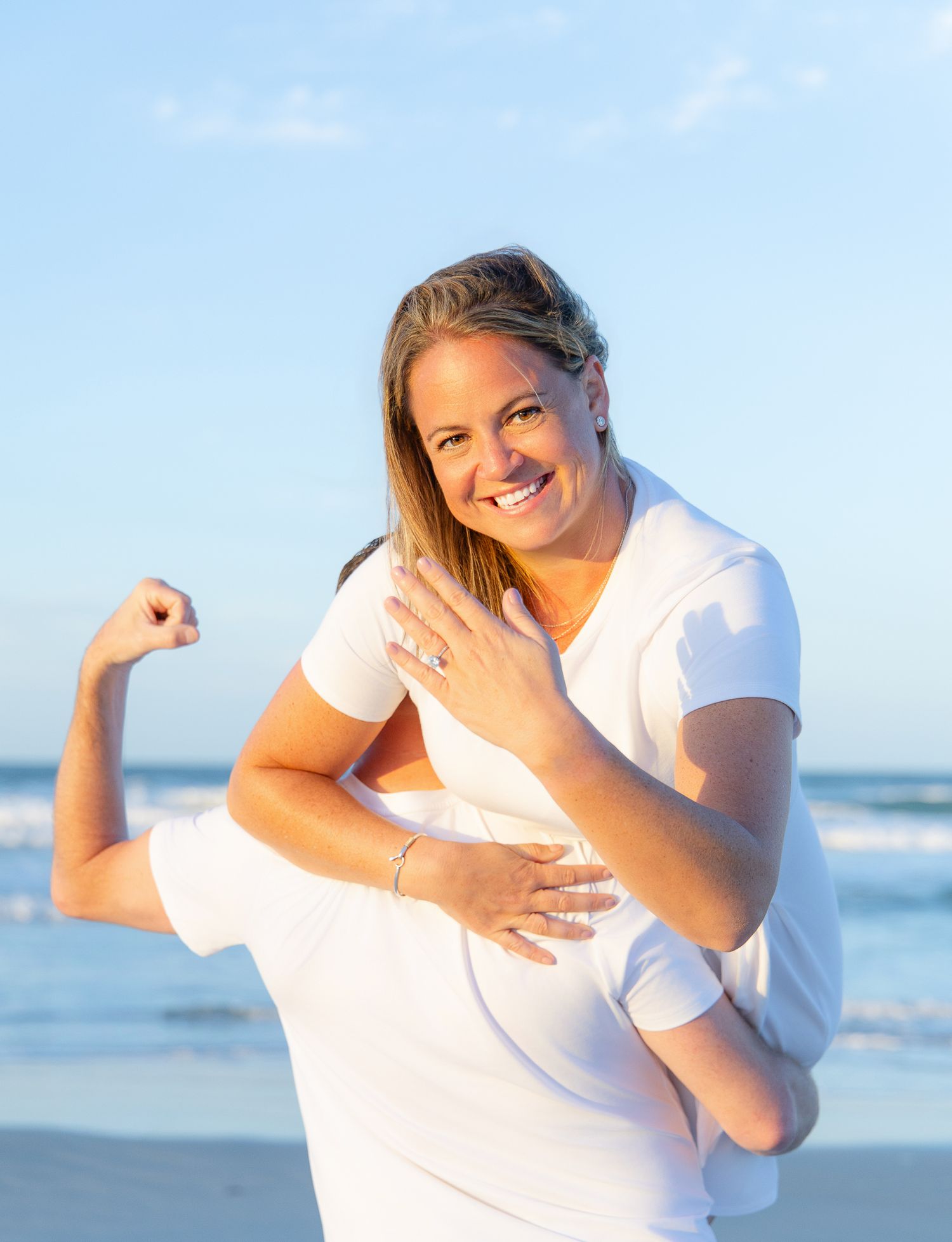 A smiling woman in a white t-shirt flexes her bicep on a sunny beach with ocean waves in the background.