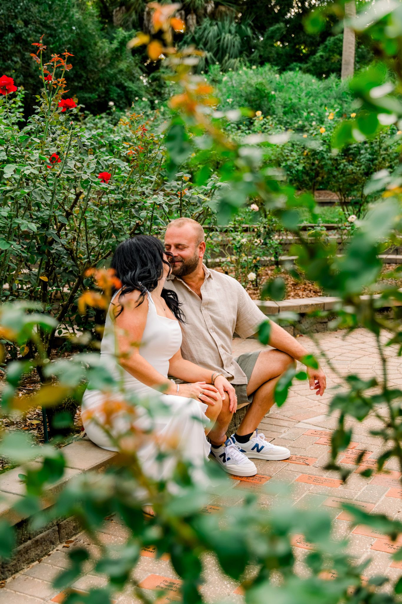 Couple sitting together on brick steps in a lush garden, surrounded by red roses and green foliage.