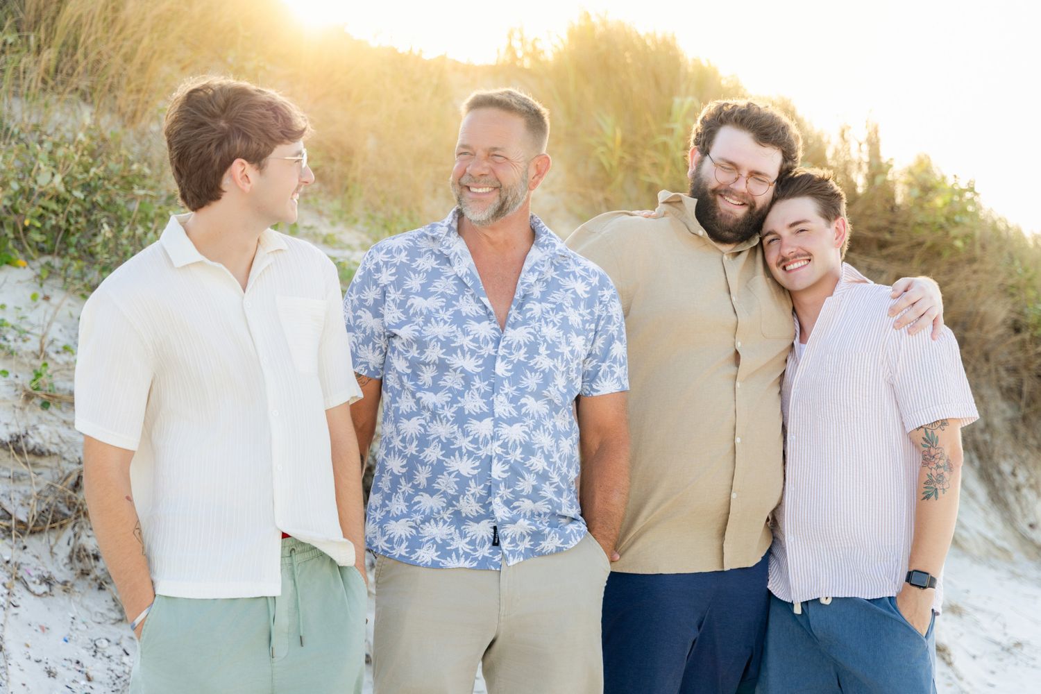 Four men smiling and embracing on a sandy beach at golden hour.