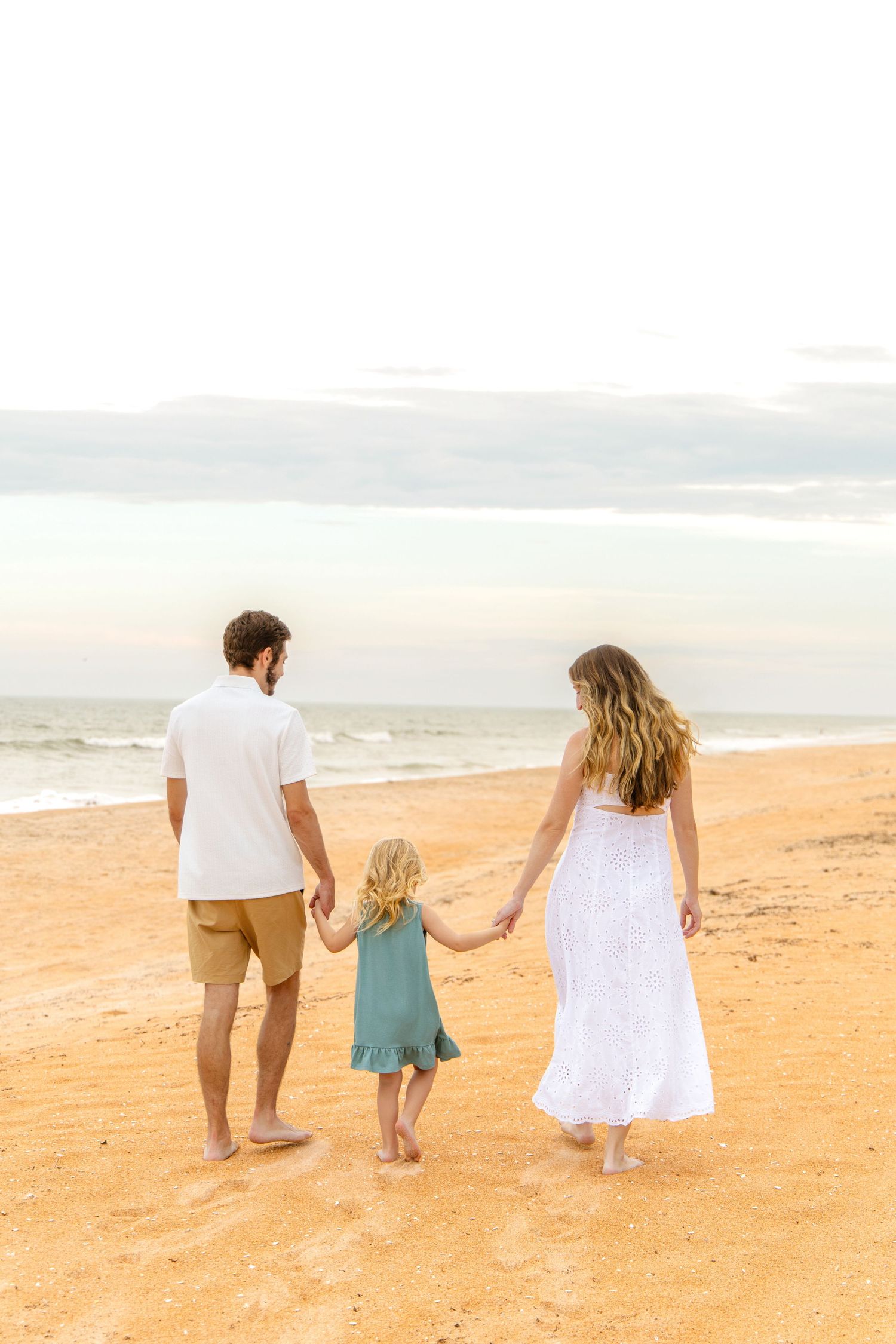 Family of three walking hand-in-hand on a sandy beach at sunset, parents in white and khaki, child in teal dress.