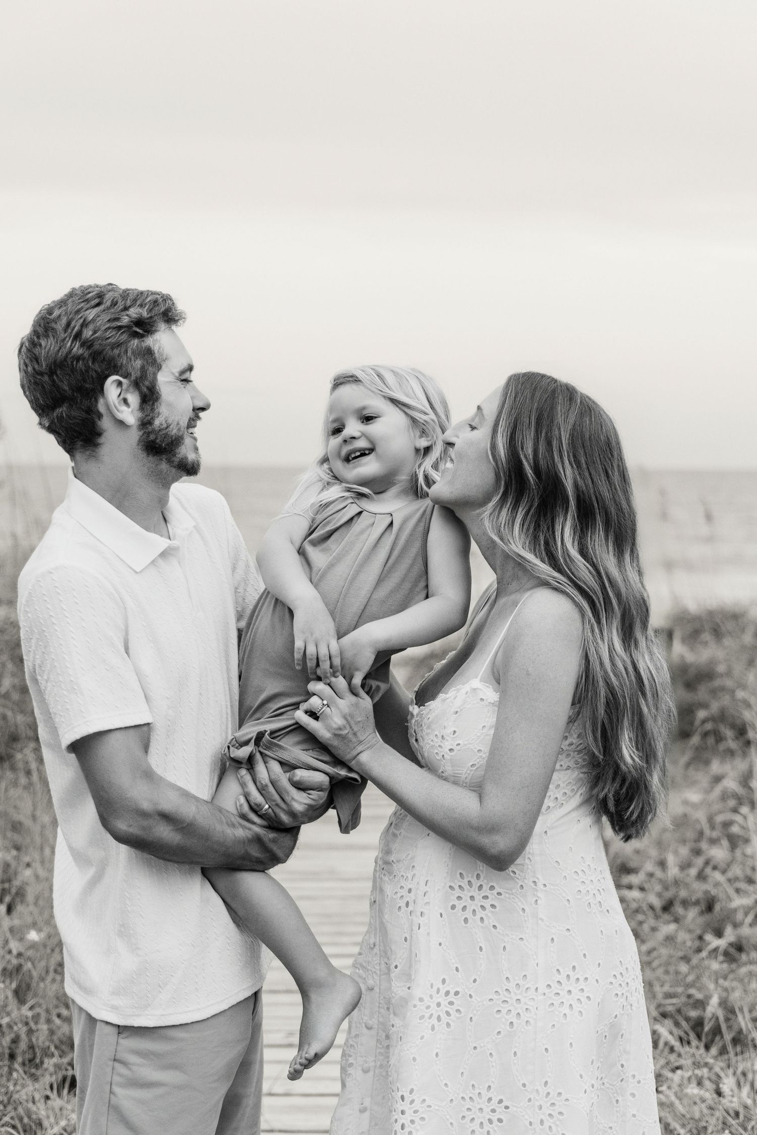 Black and white photo of a couple at the beach, man holding toddler while woman kisses child on cheek.