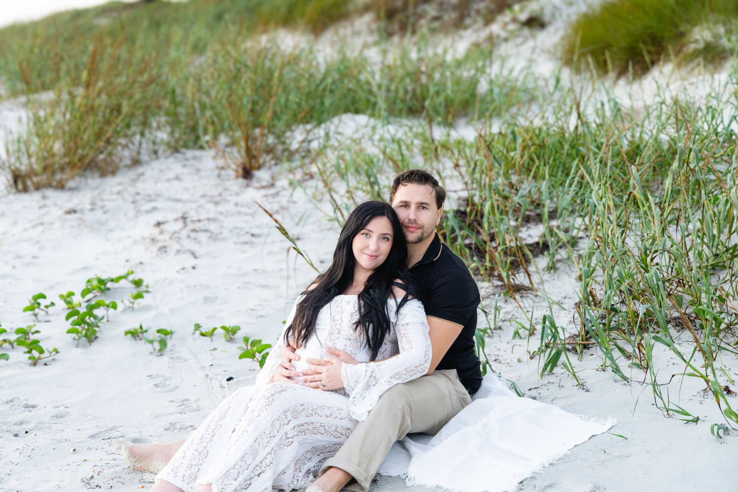 Couple sitting together on white sand dunes with beach grass, woman in lace dress, man in black polo shirt.