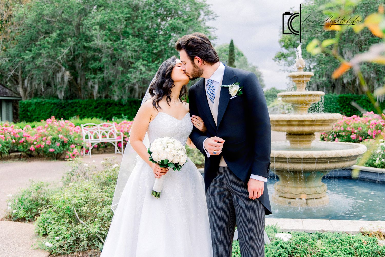 Bride and groom share a kiss near an elegant stone fountain surrounded by blooming roses in a garden.