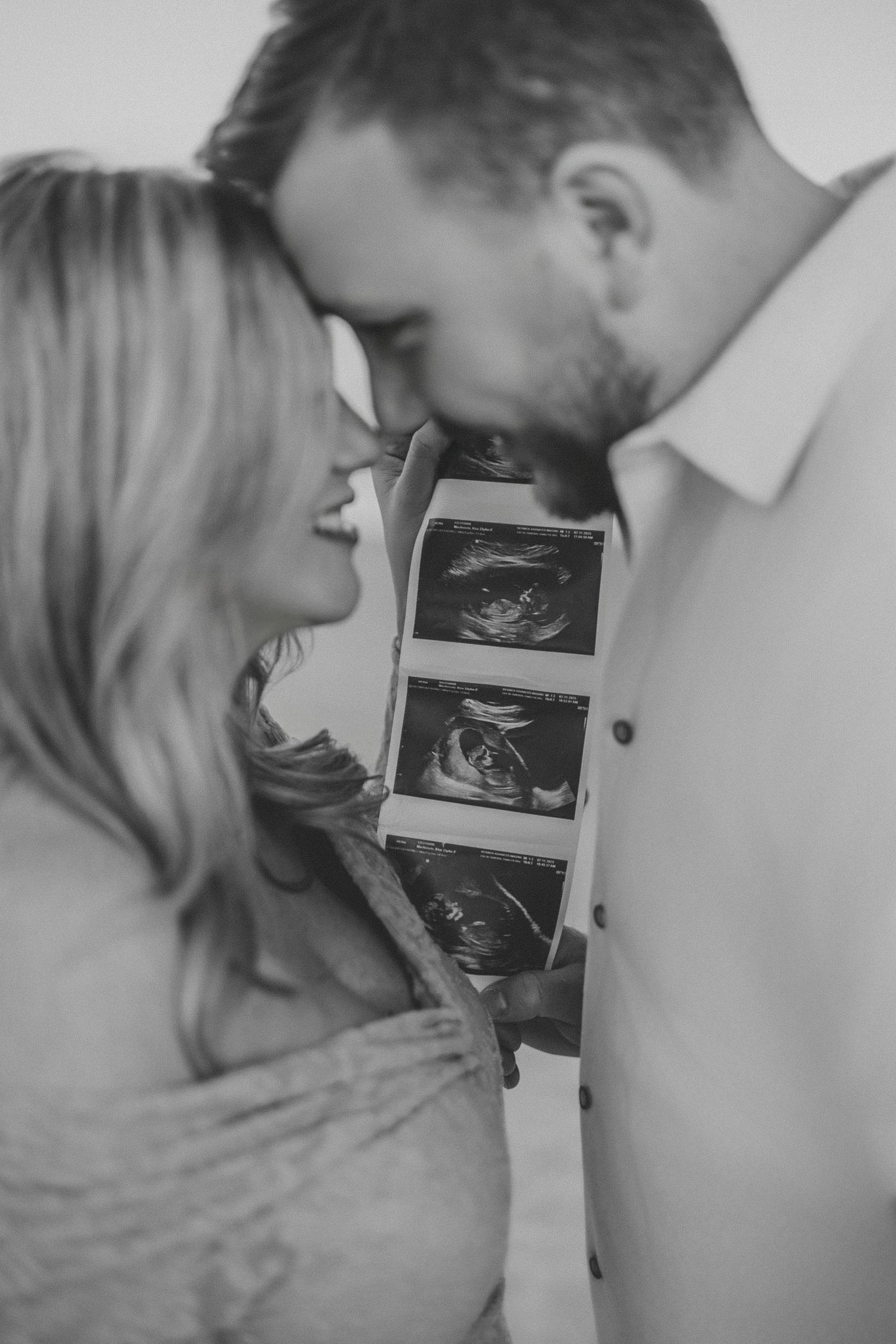 A couple shares an intimate moment while holding ultrasound photos in a black and white portrait.