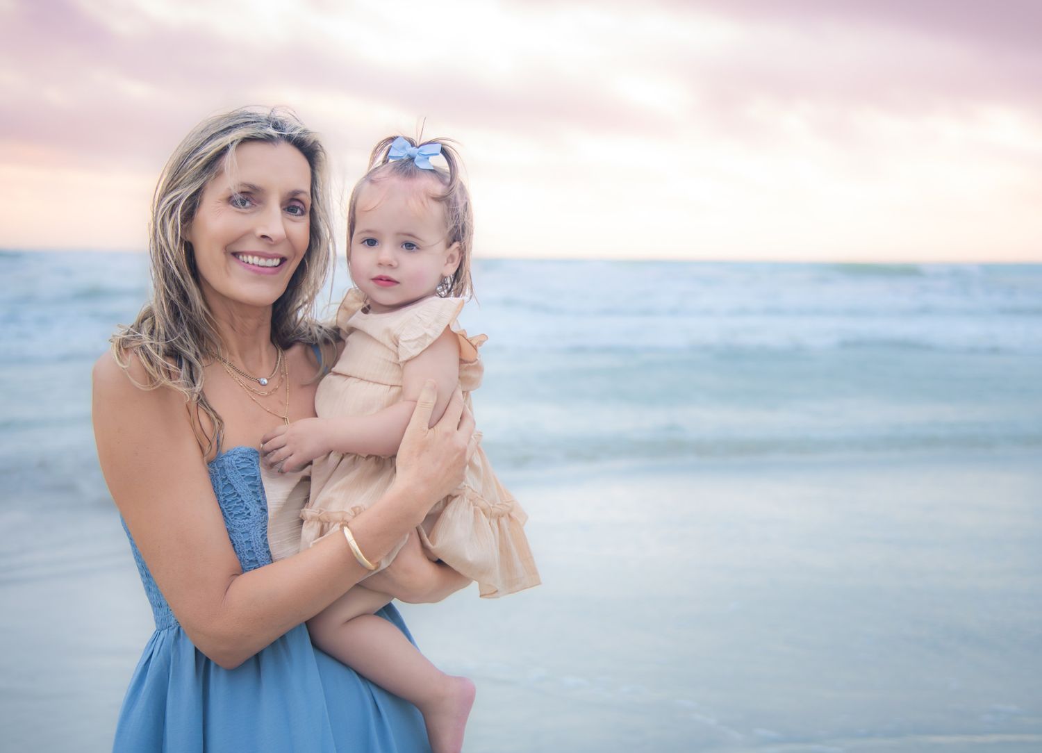Woman in blue dress holds baby girl on beach at sunset, ocean waves in background.