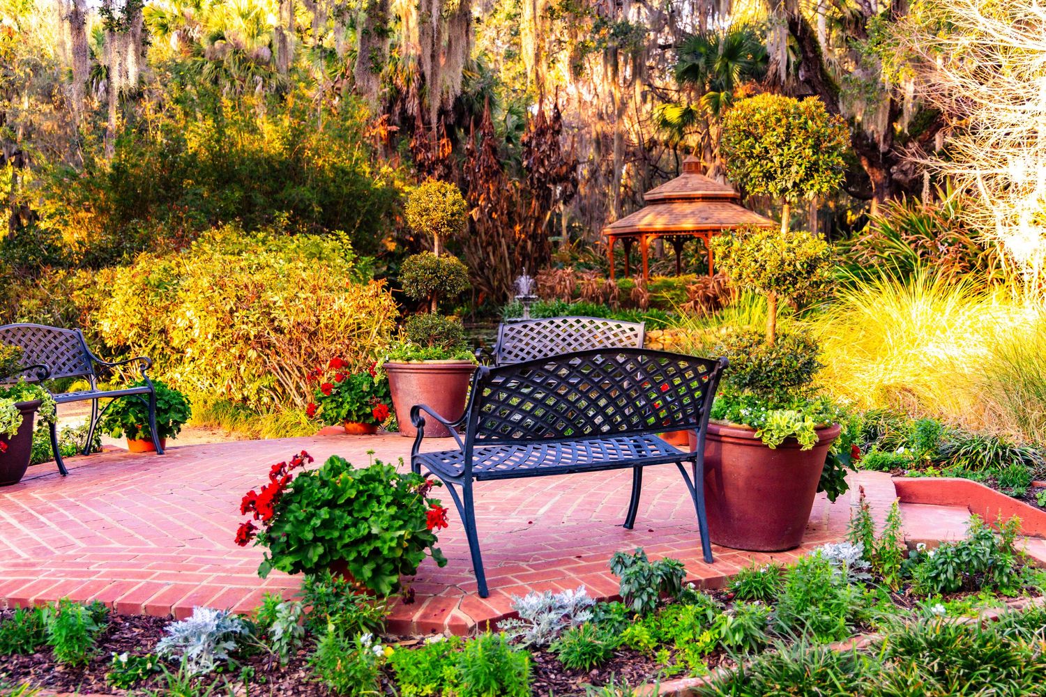 A lush garden patio with brick pavers, colorful potted plants, and an umbrella surrounded by natural greenery in golden light.