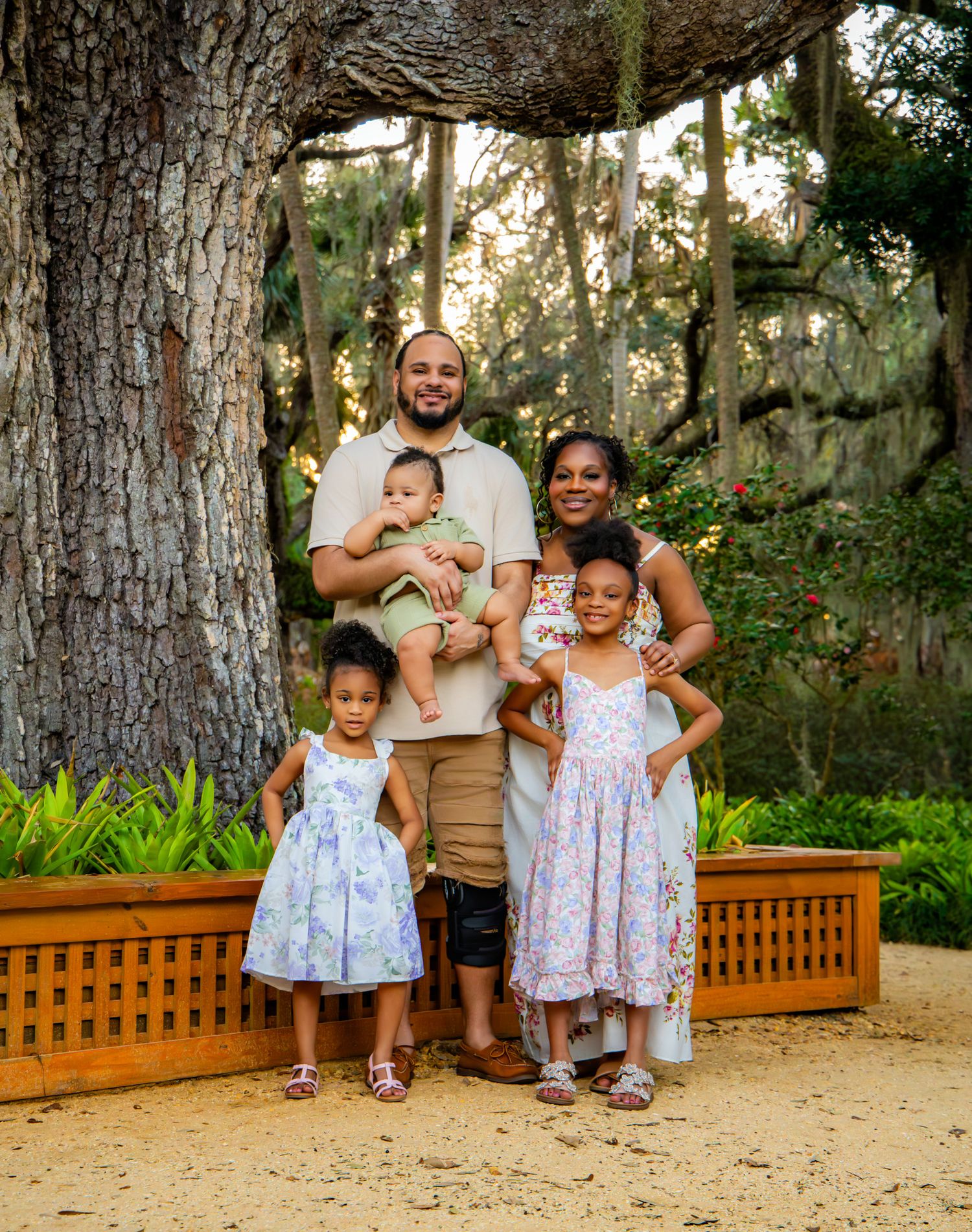 A family stands together in front of a large oak tree with Spanish moss on a sunny afternoon in a garden setting.