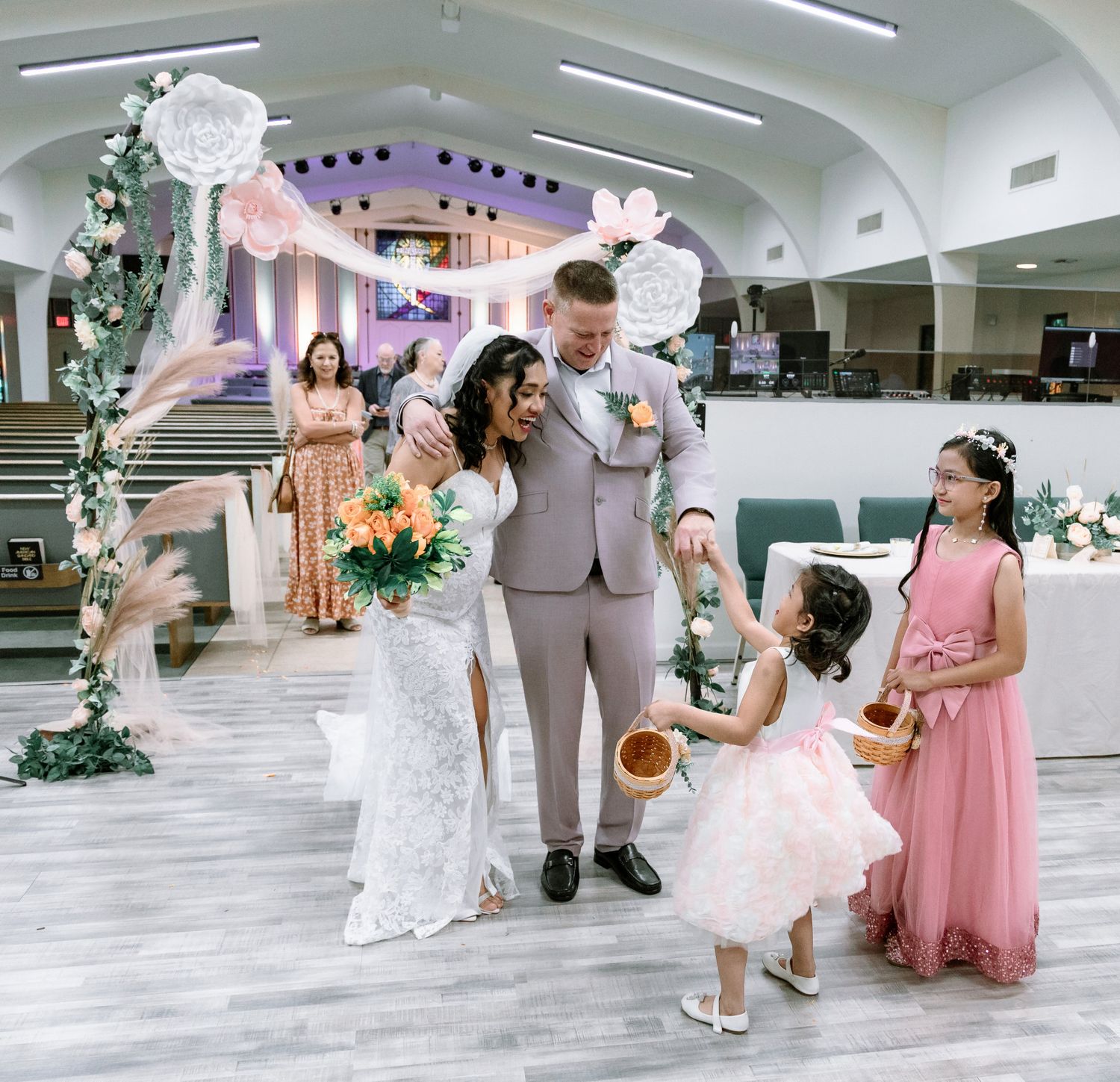 Bride and groom celebrate with flower girls at church wedding ceremony, surrounded by floral arch decorations.