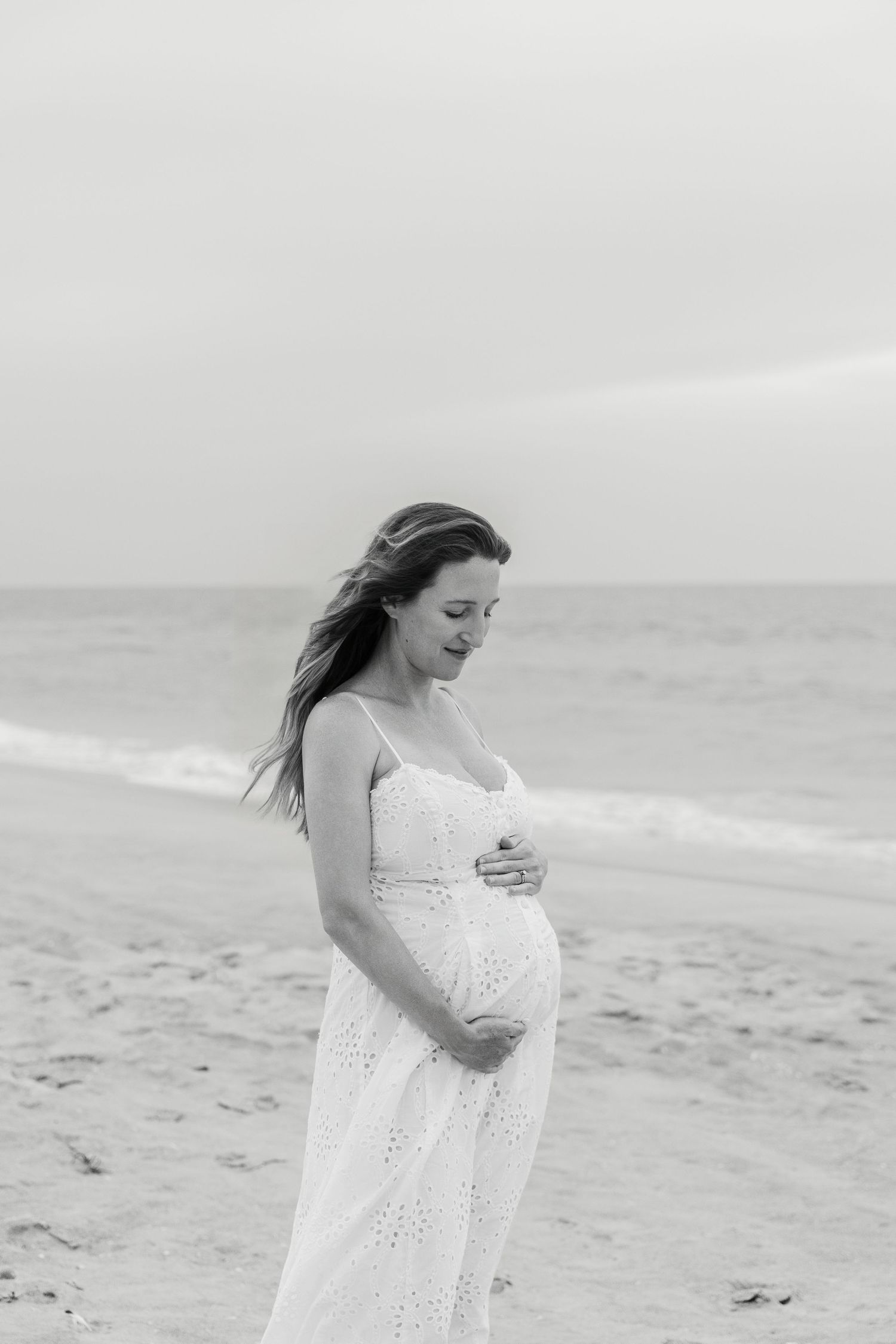 Black and white photo of a pregnant woman in a white dress cradling her belly on a beach.