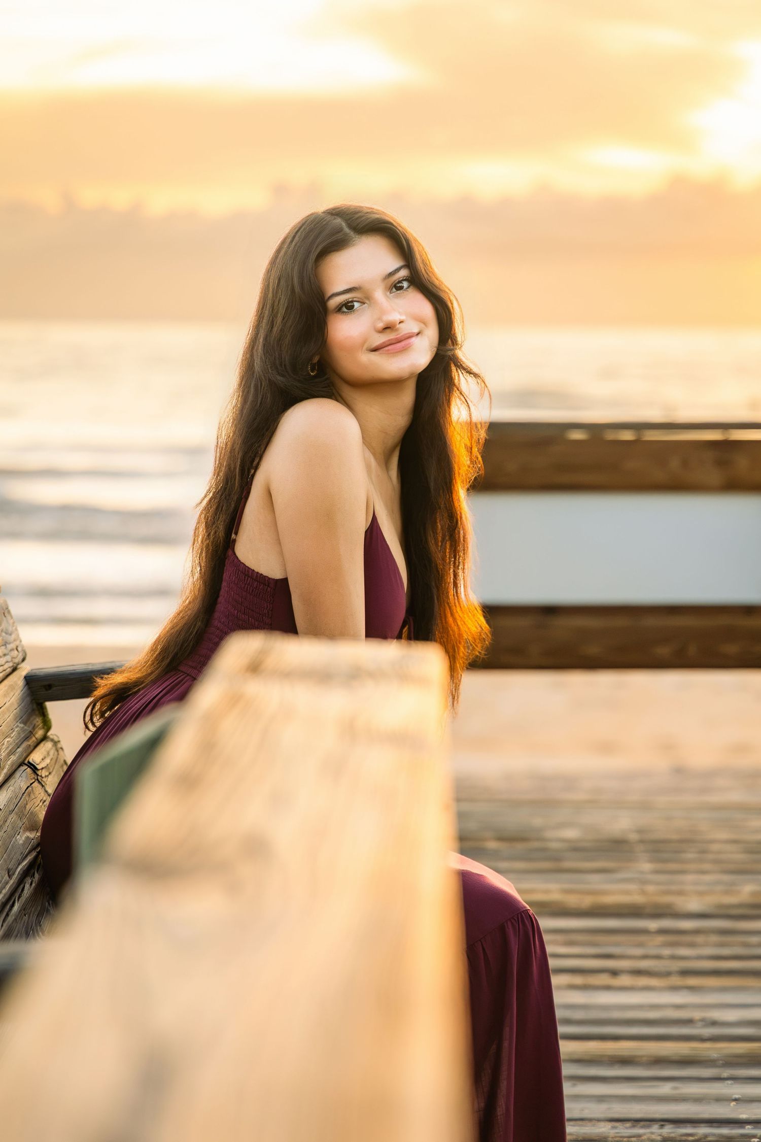 Young woman in burgundy dress sitting on wooden pier at golden sunset by the ocean, smiling over shoulder.