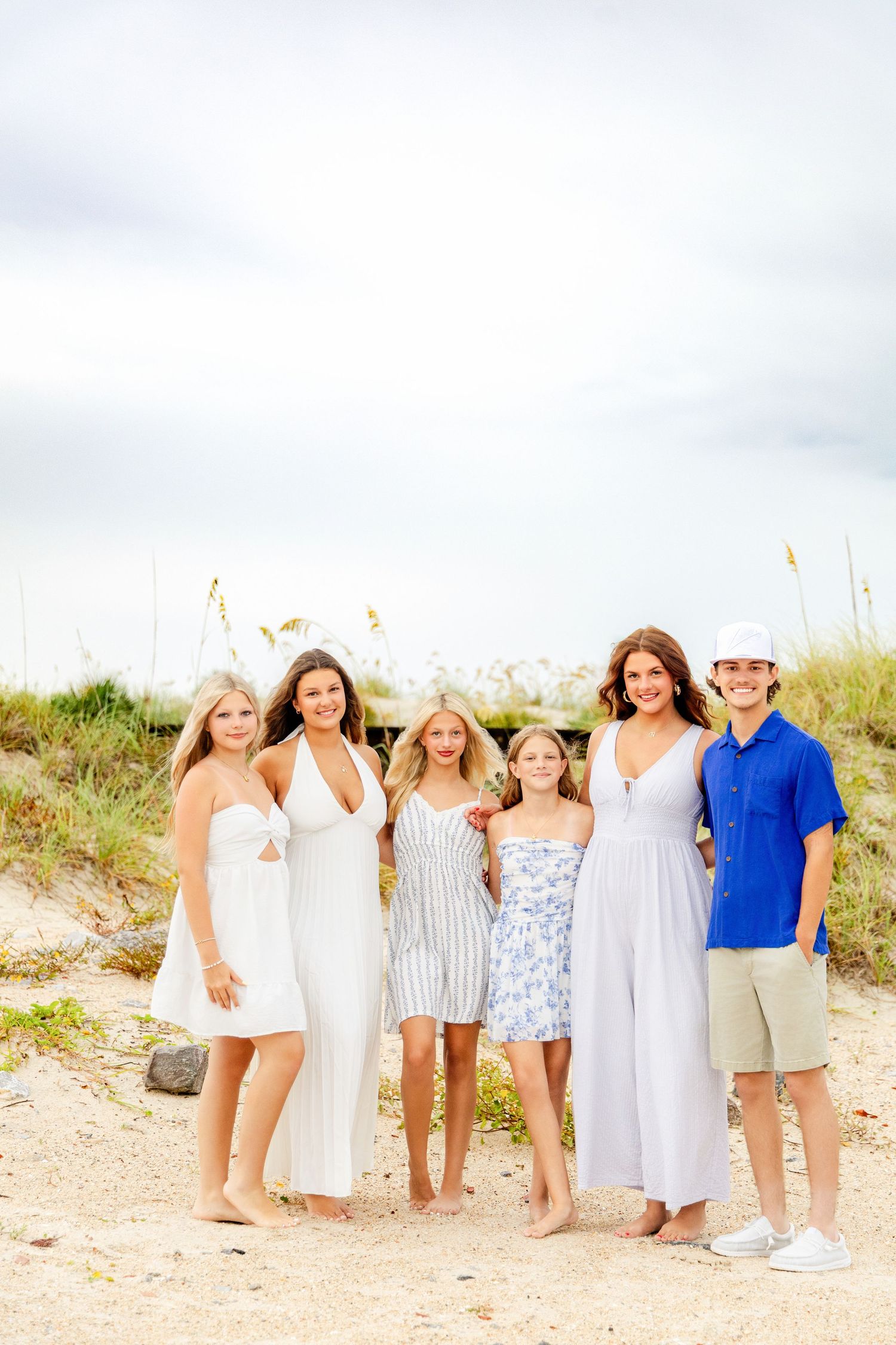 A family of six poses together on a sandy beach, wearing white, blue, and floral outfits on a cloudy day.