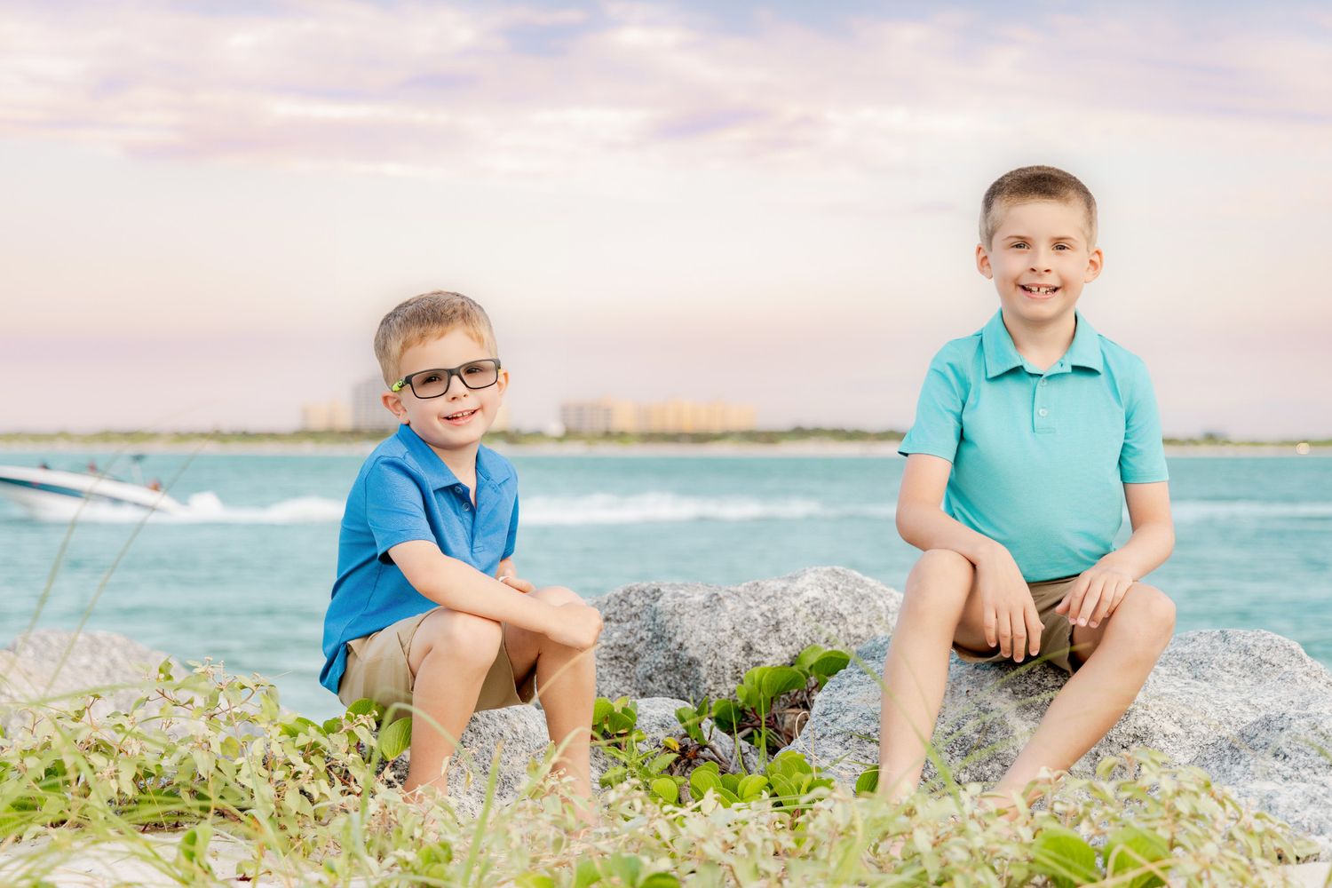 Two young boys sitting on rocks by the ocean shore, smiling at camera on a sunny beach day.