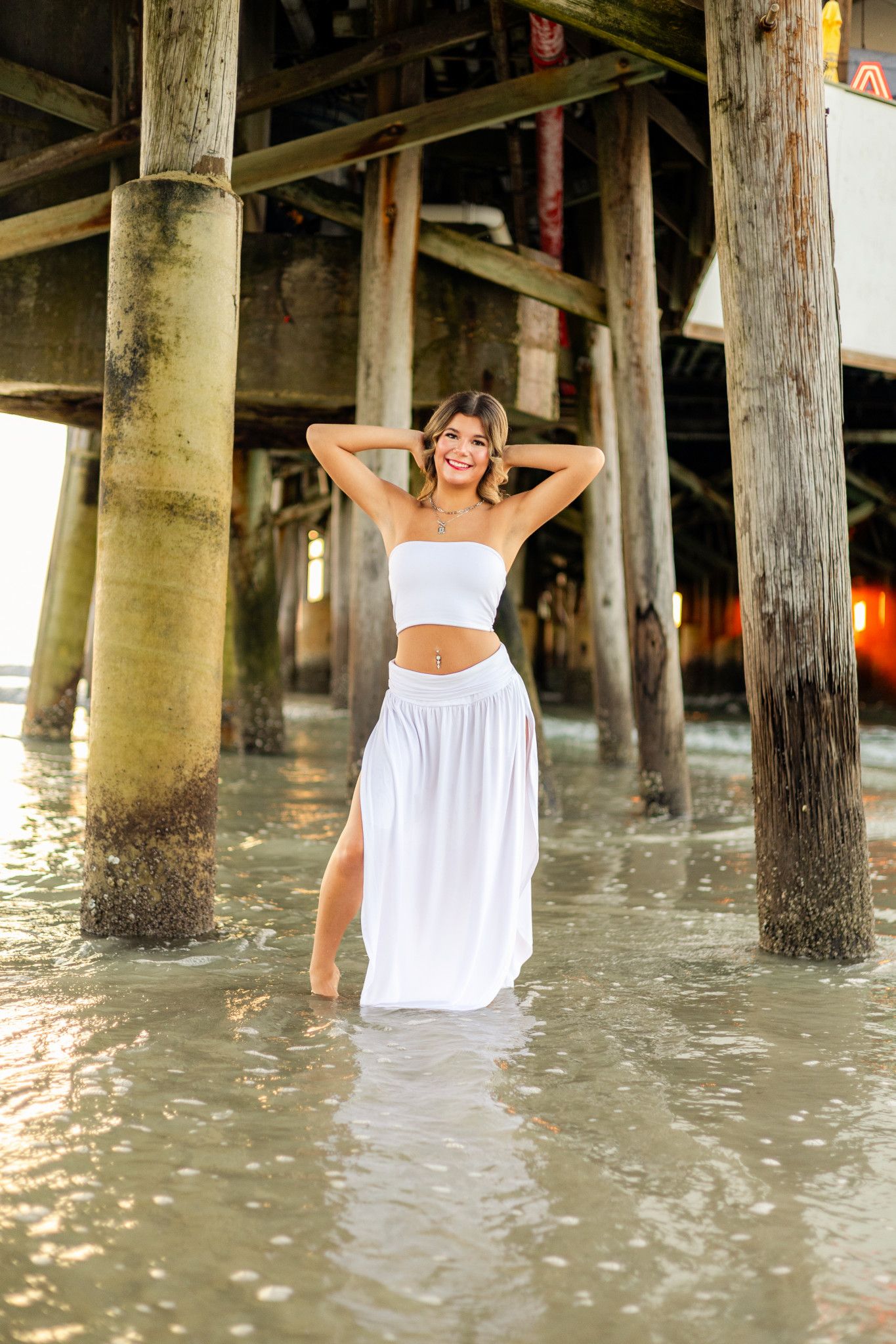 Young woman in white crop top and maxi skirt posing under a beach pier in shallow water.